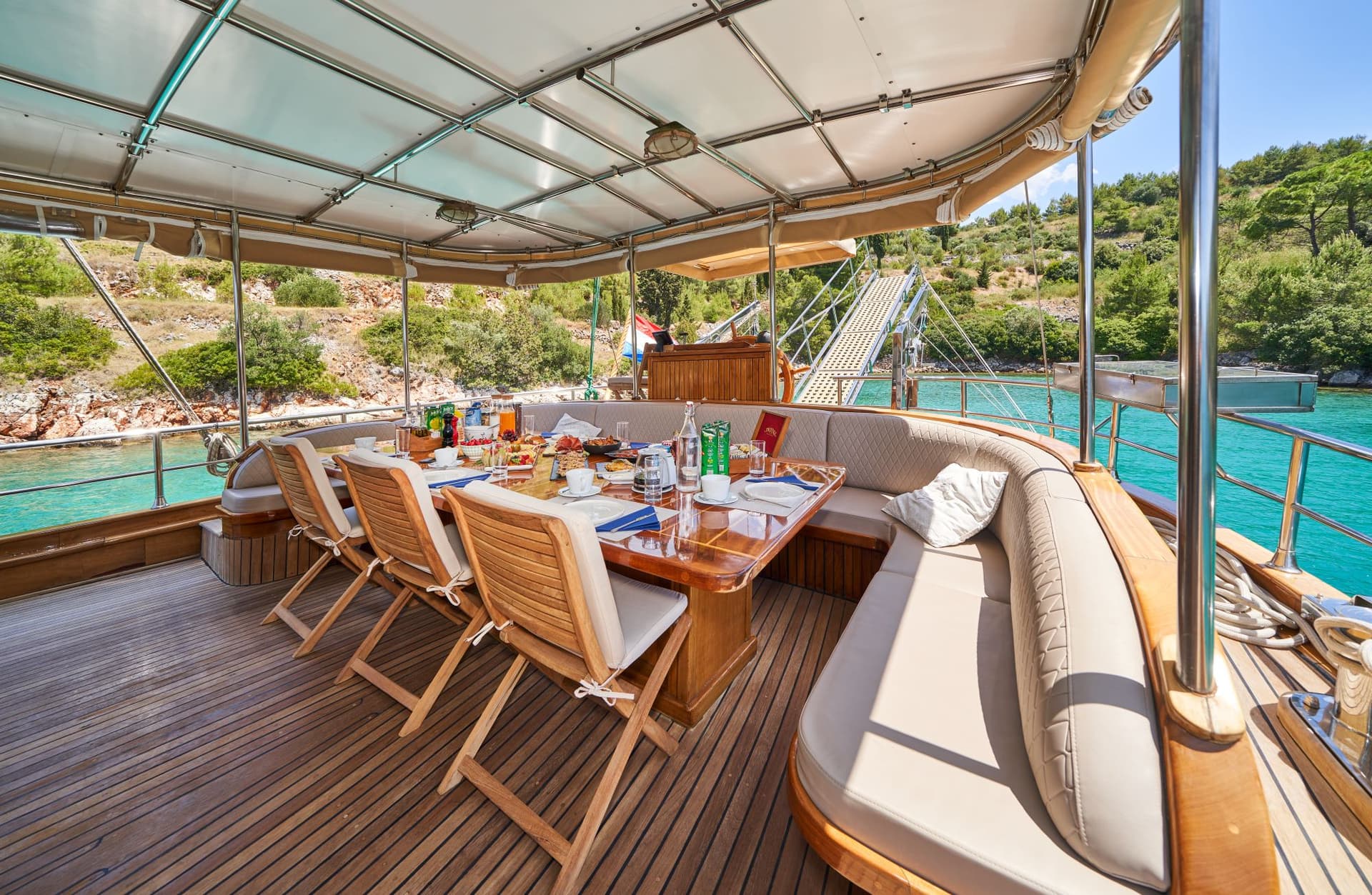 Outdoor dining area on a yacht with breakfast spread near a lush, rocky coastline.