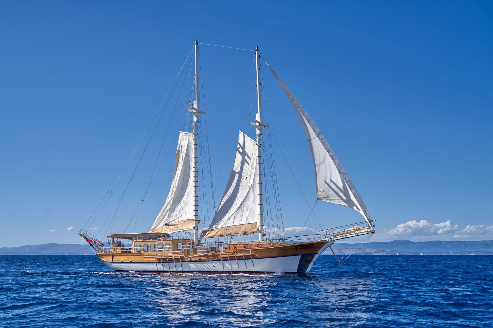 Wooden sailboat with white sails on deep blue sea under clear blue sky near coast.