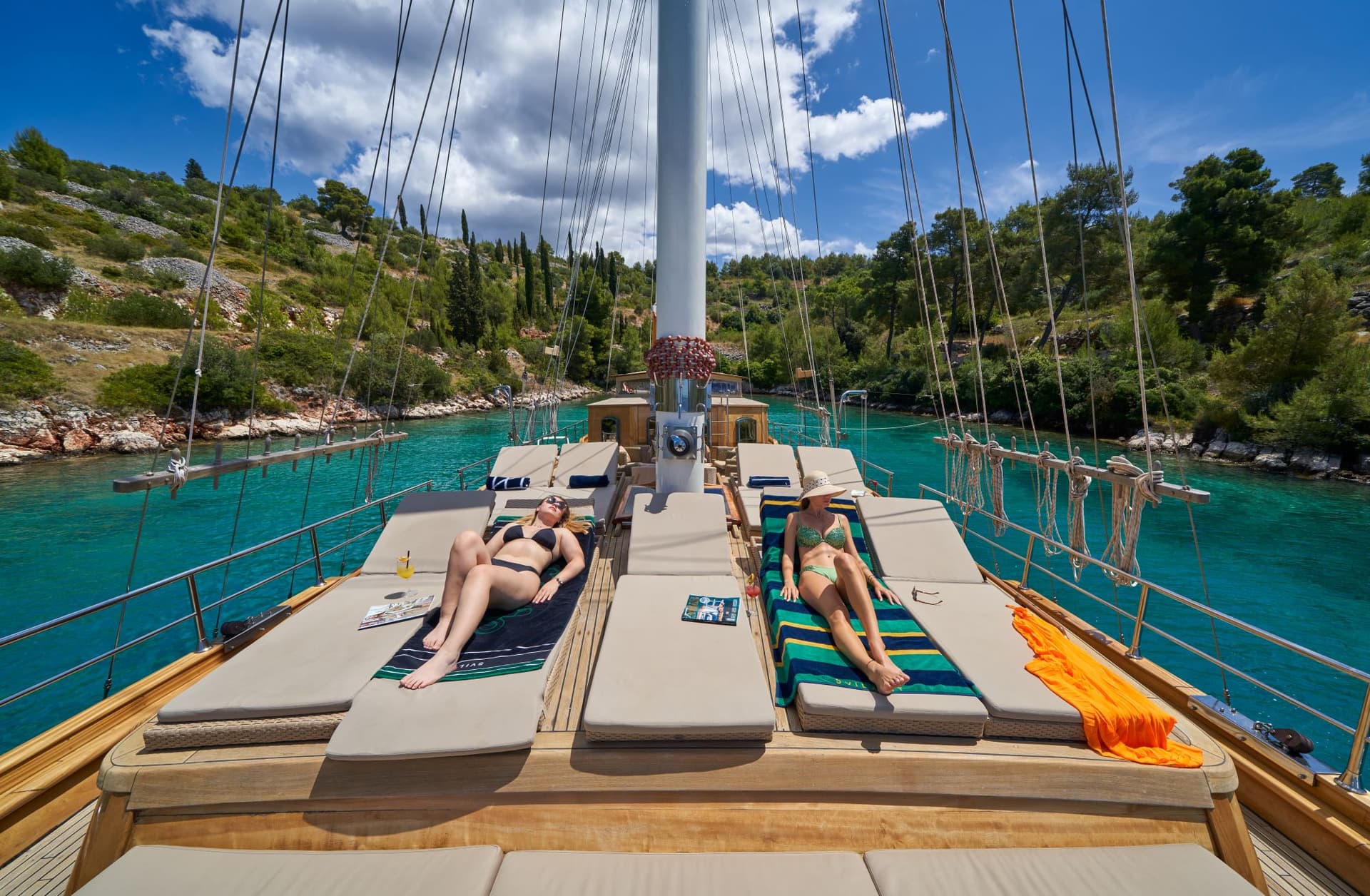 Two women sunbathing on deck of wooden boat in turquoise cove with green hills