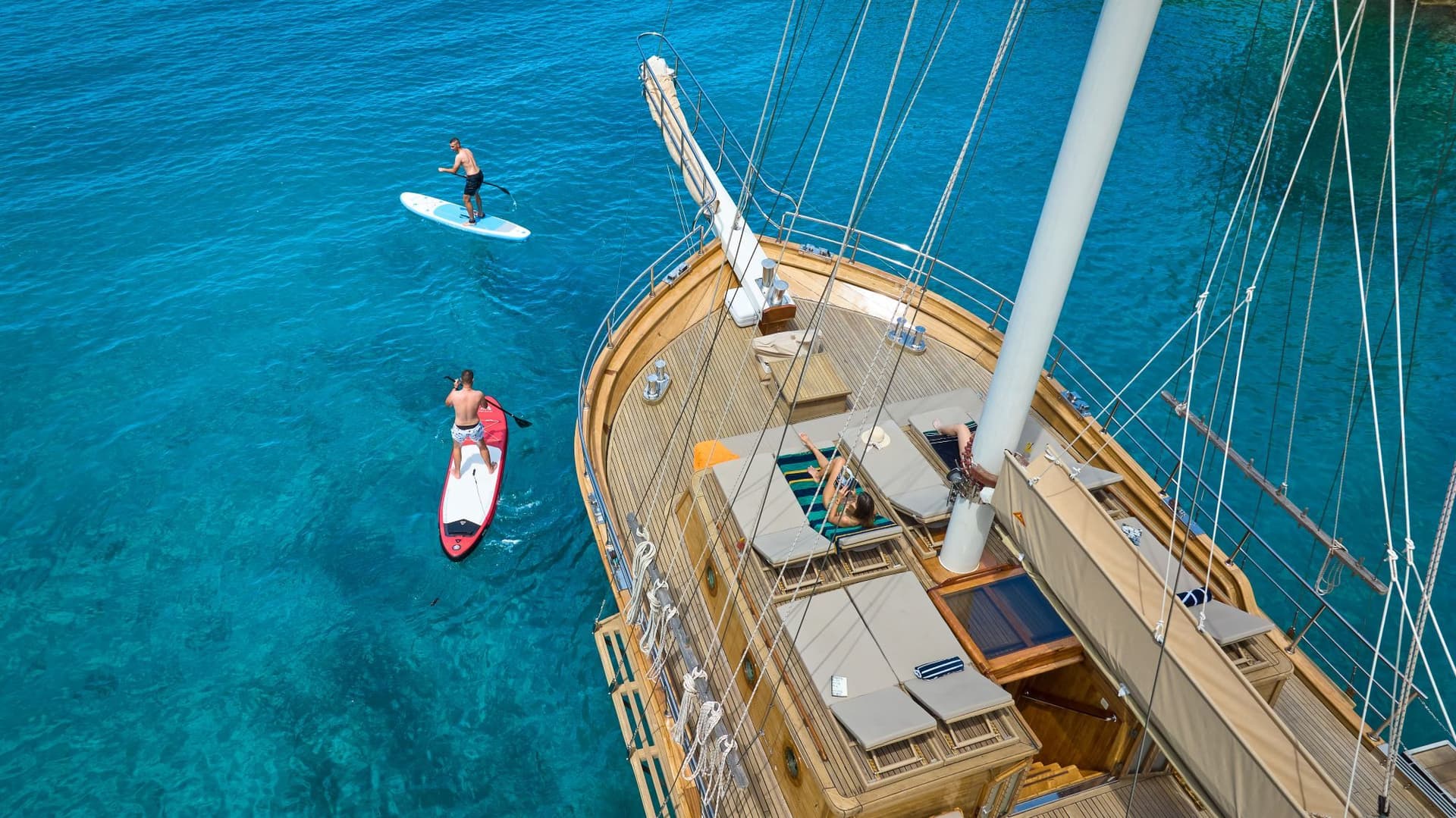 Paddleboarding near a wooden yacht deck with people sunbathing on turquoise water.