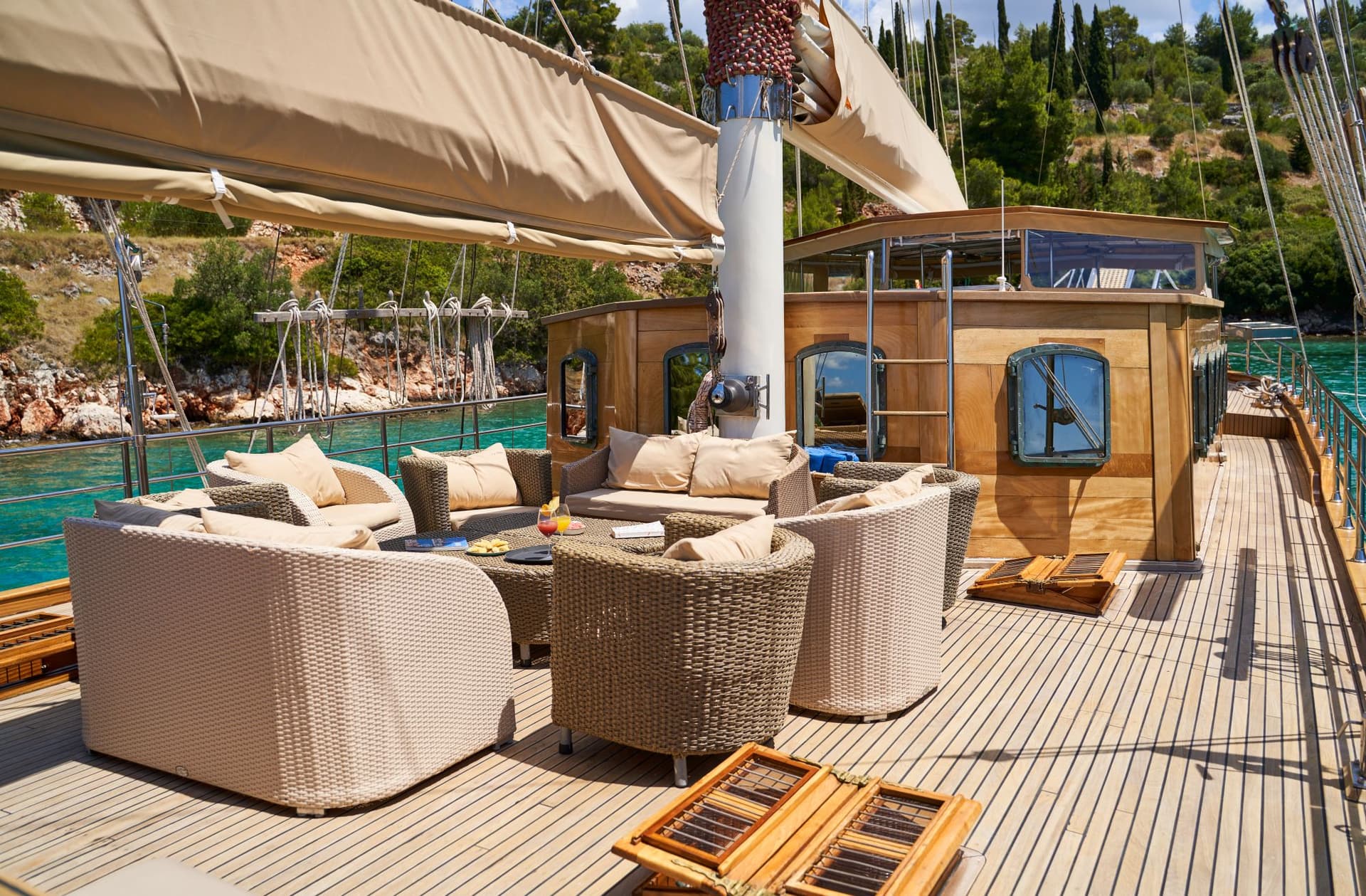 Sundeck lounge area with wicker furniture on a wooden boat near a lush green coastline.