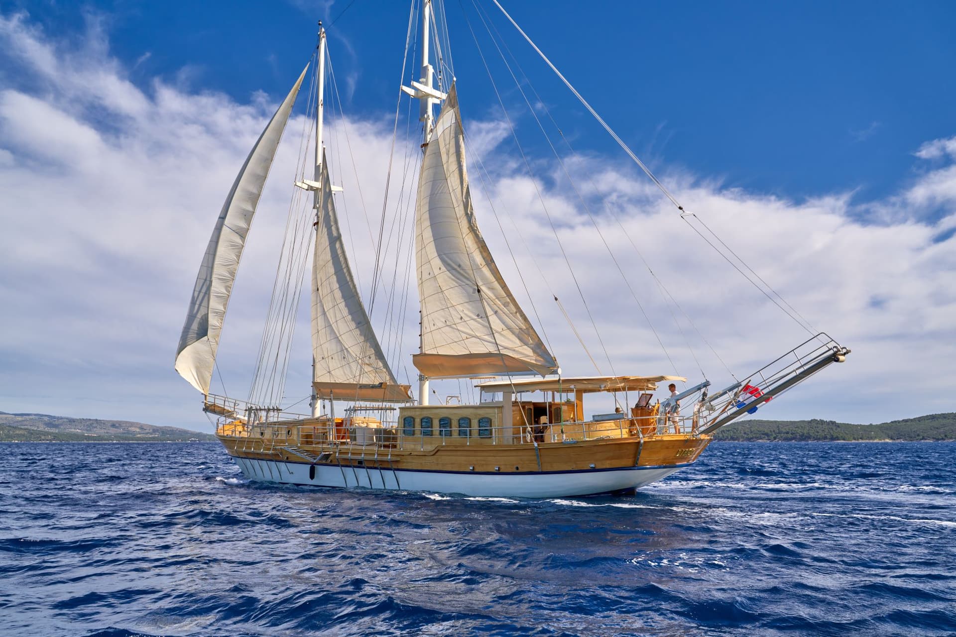 Wooden sailboat with open sails on deep blue sea near a green coastline under a cloudy sky.