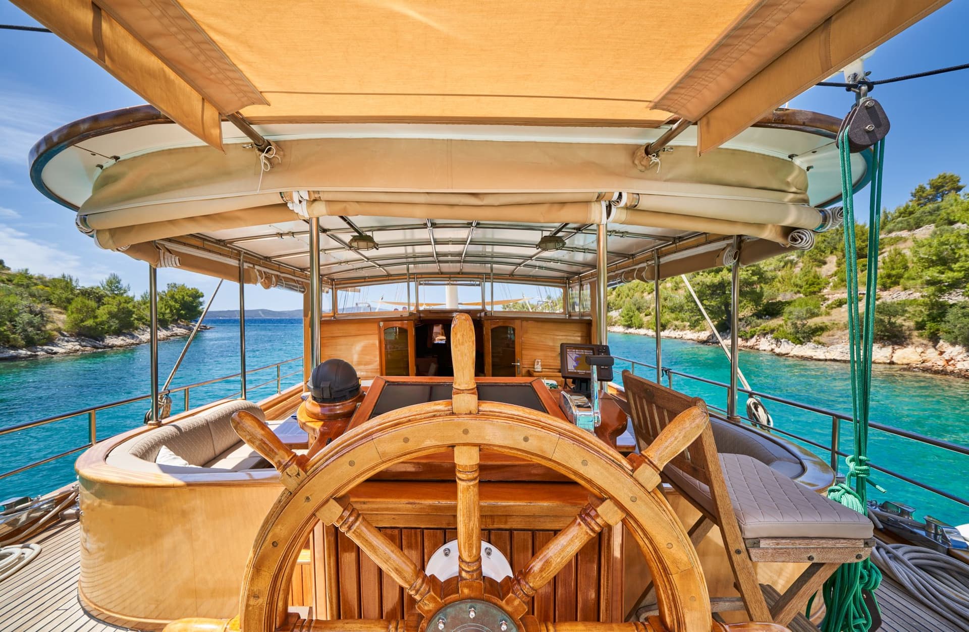 Wooden boat helm on deck navigating turquoise channel between tree-covered shores