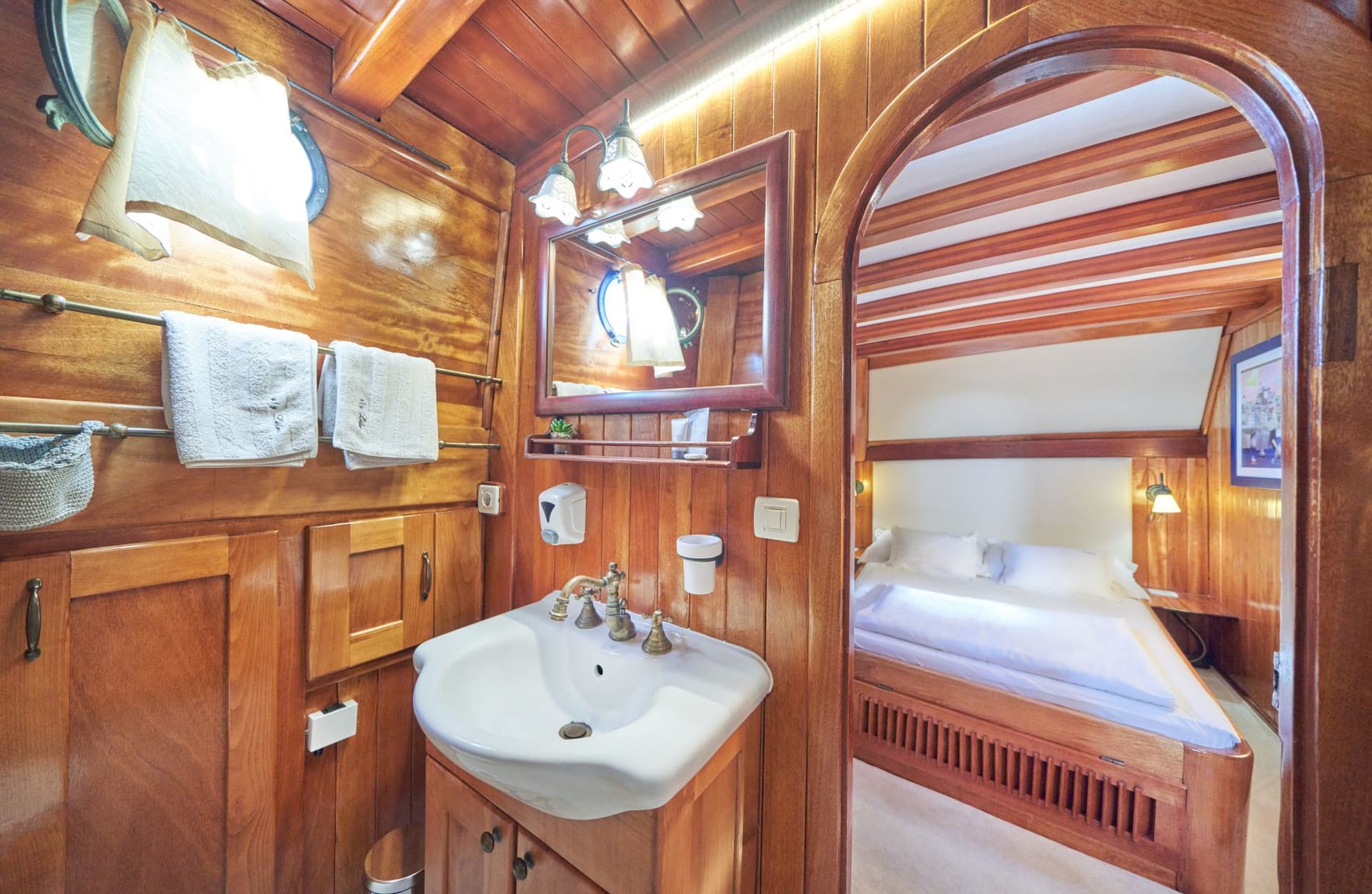 Wood-paneled boat bathroom with white sink, mirror, and view into adjacent cabin with bed.