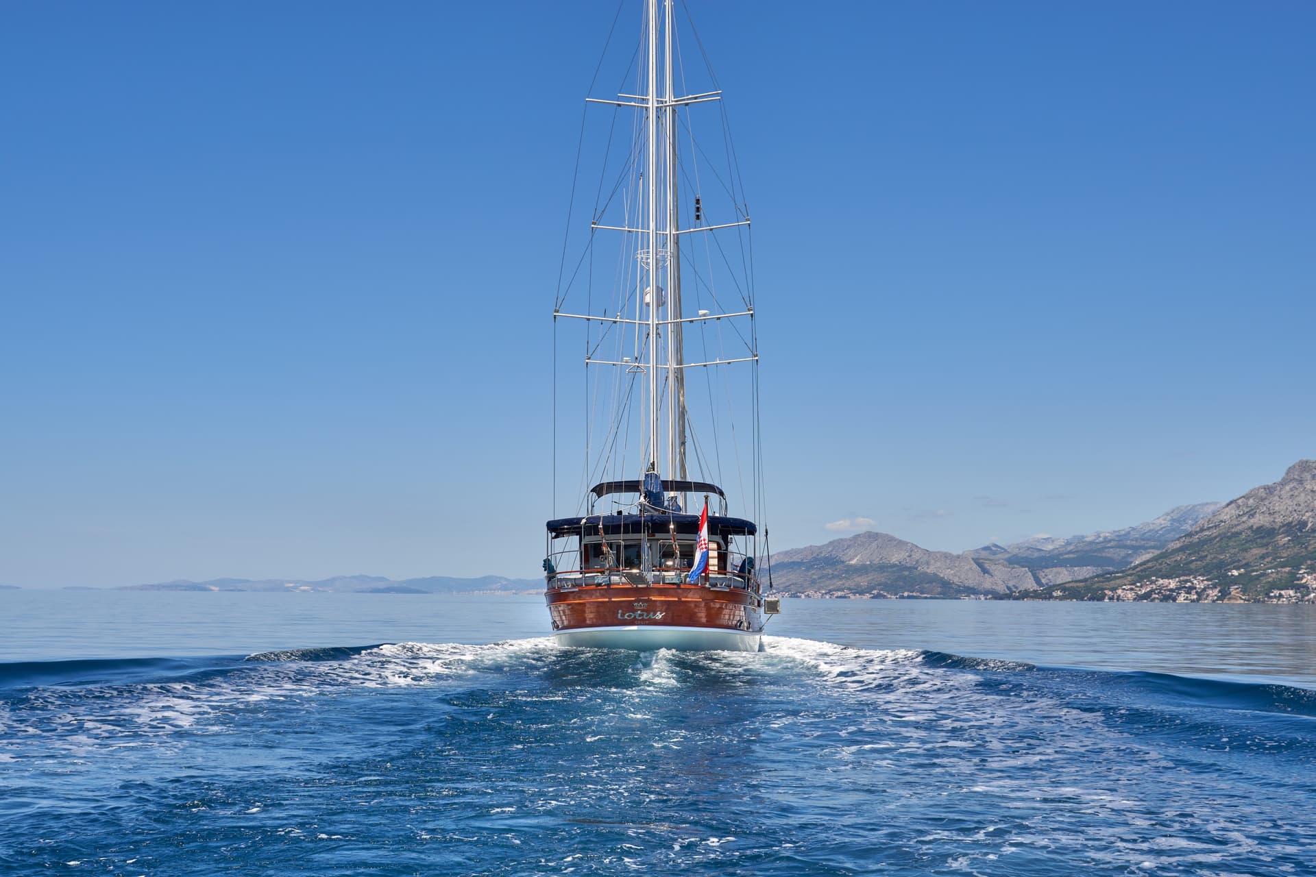 Wooden sailboat named Lotus leaving a wake on blue sea near rocky coastline.