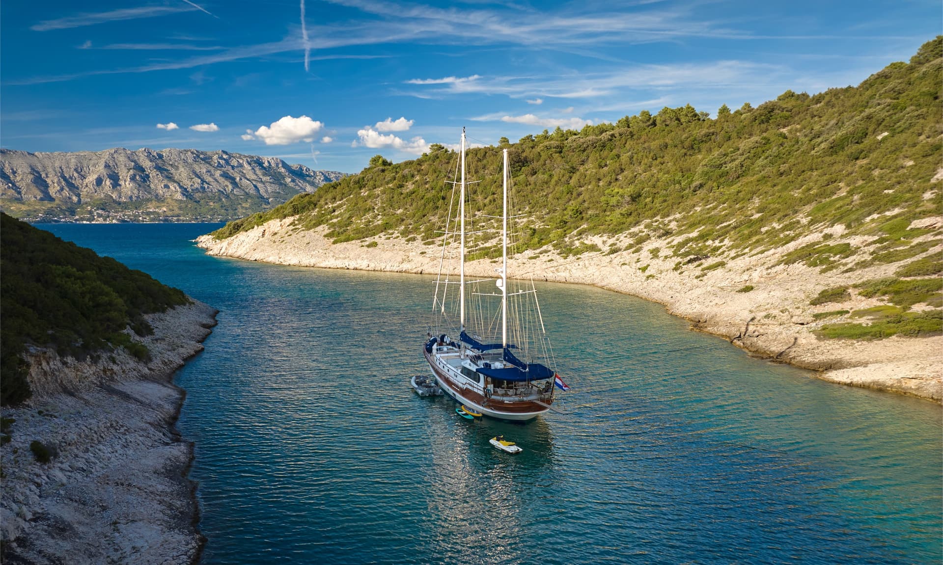 Large sailboat anchored in a cove with steep, green, rocky hillsides under a blue sky.