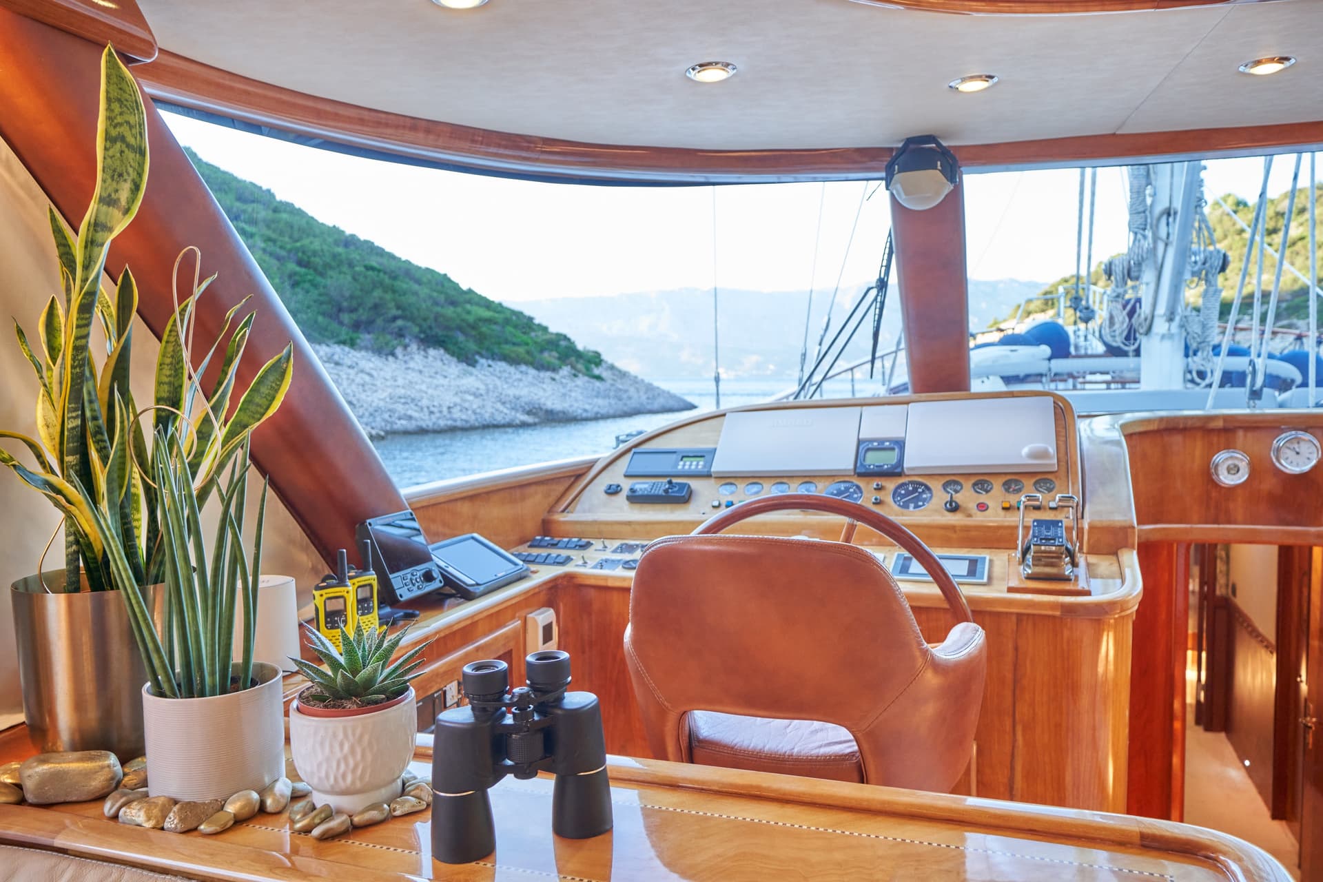 Yacht helm station with wood trim, binoculars, and plants overlooking a green, rocky coastline.