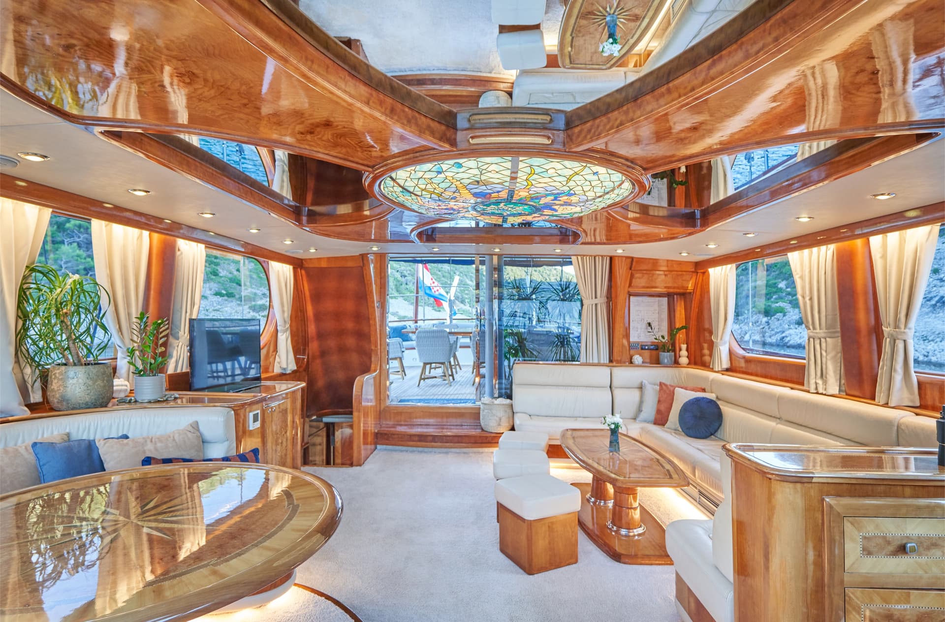Luxury yacht salon with glossy wood paneling, white leather seating, and view of rocky coastline.