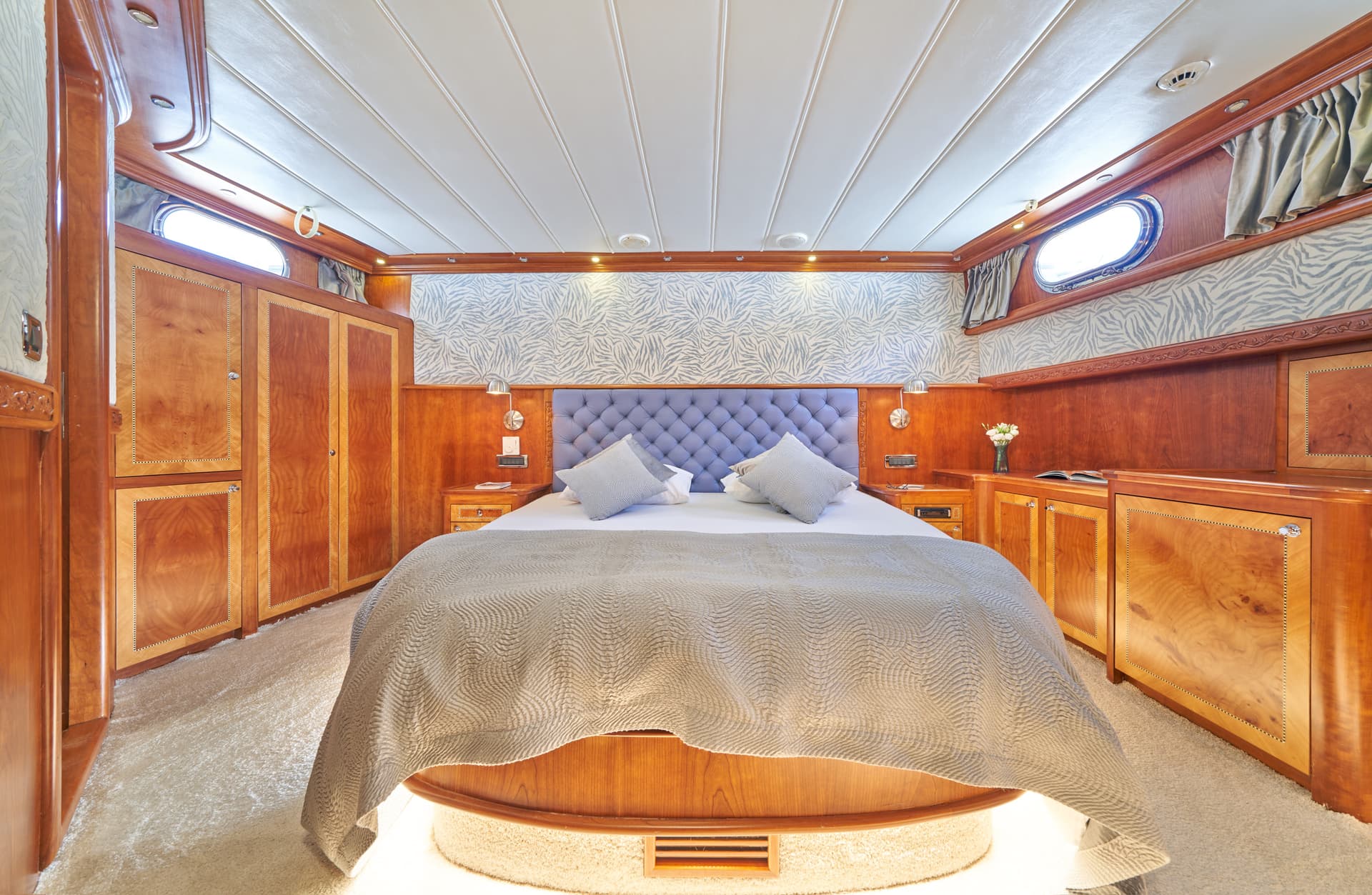 Bedroom cabin with wood paneling, tufted headboard, and oval porthole windows on a boat.