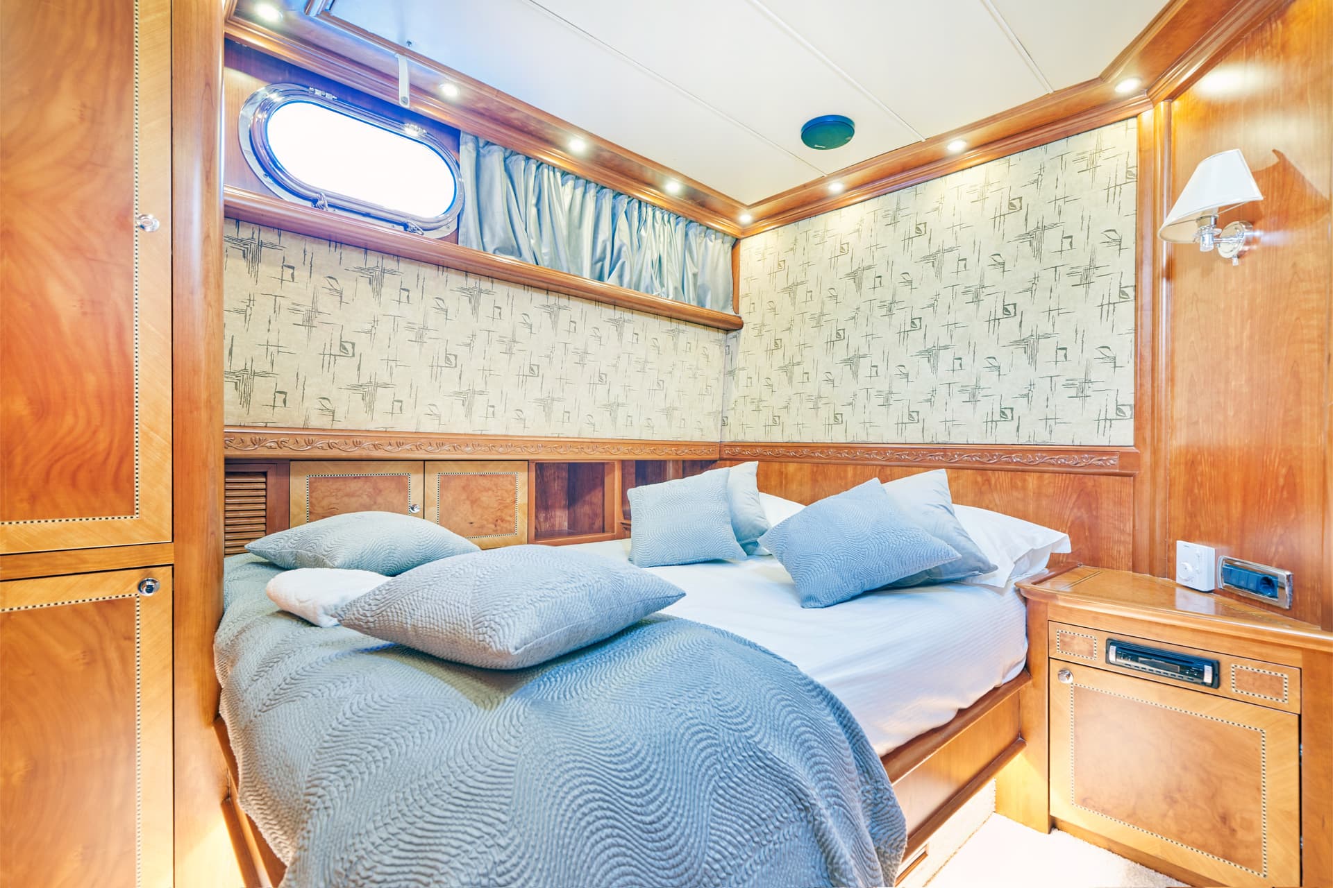 Cabin bedroom with wood paneling, oval porthole window, and blue bedding on a boat.