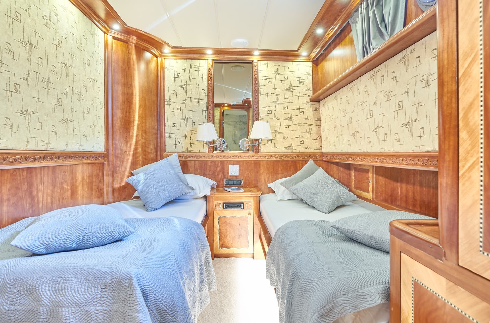 Twin cabin interior with polished wood paneling, patterned wallpaper, and light blue bedding.