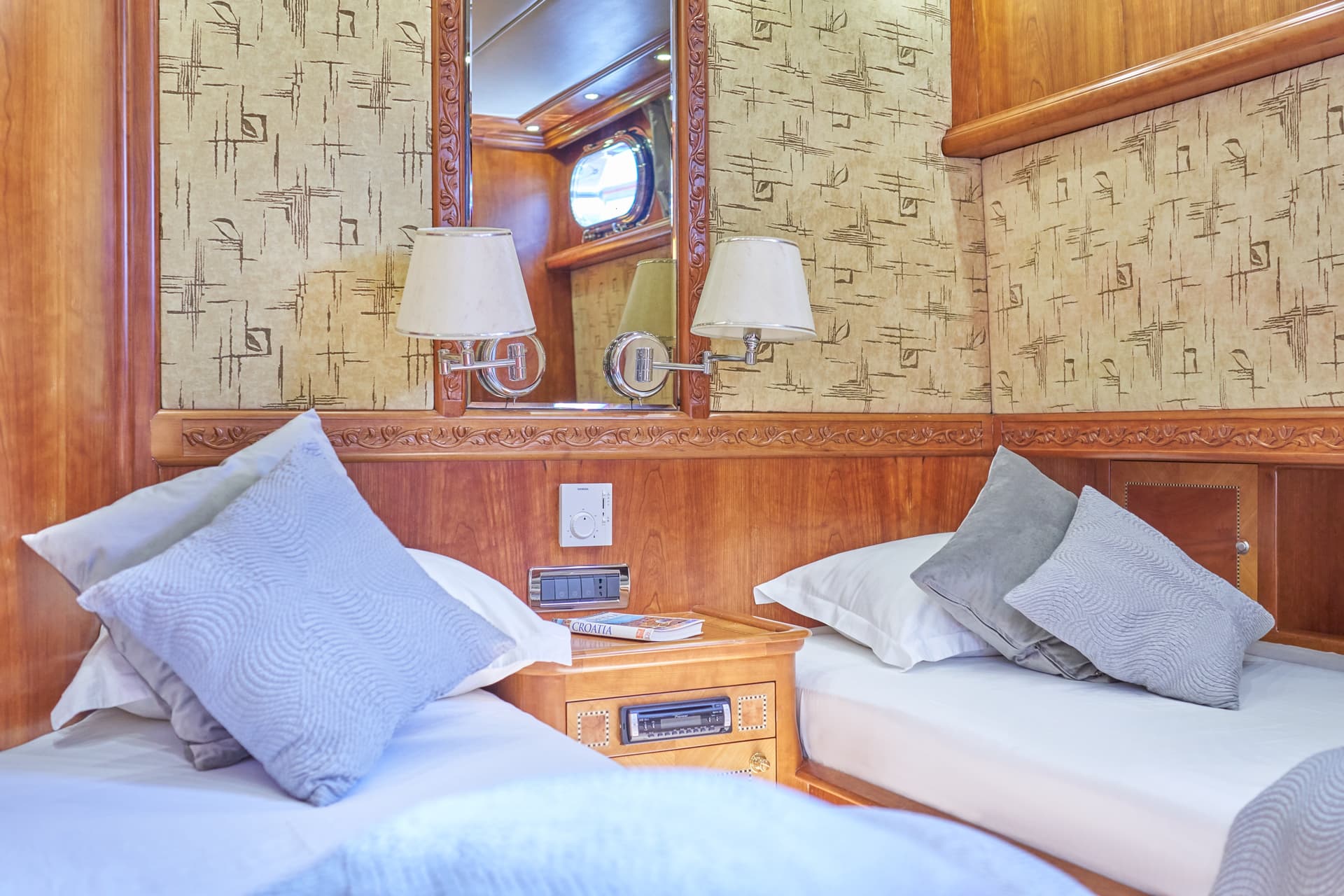 Twin beds in cabin with wood paneling, patterned wallpaper, and a mirror reflecting a porthole.