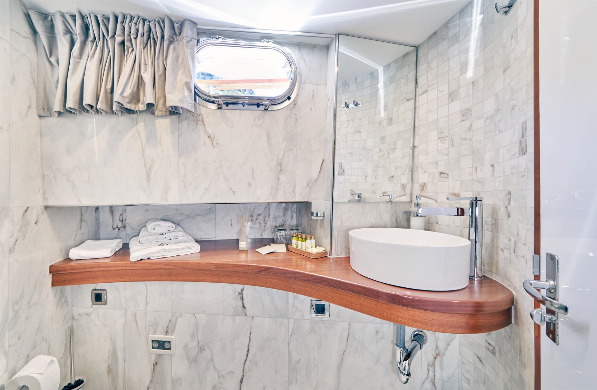 Modern yacht bathroom with marble walls, wood counter, vessel sink, and porthole window.