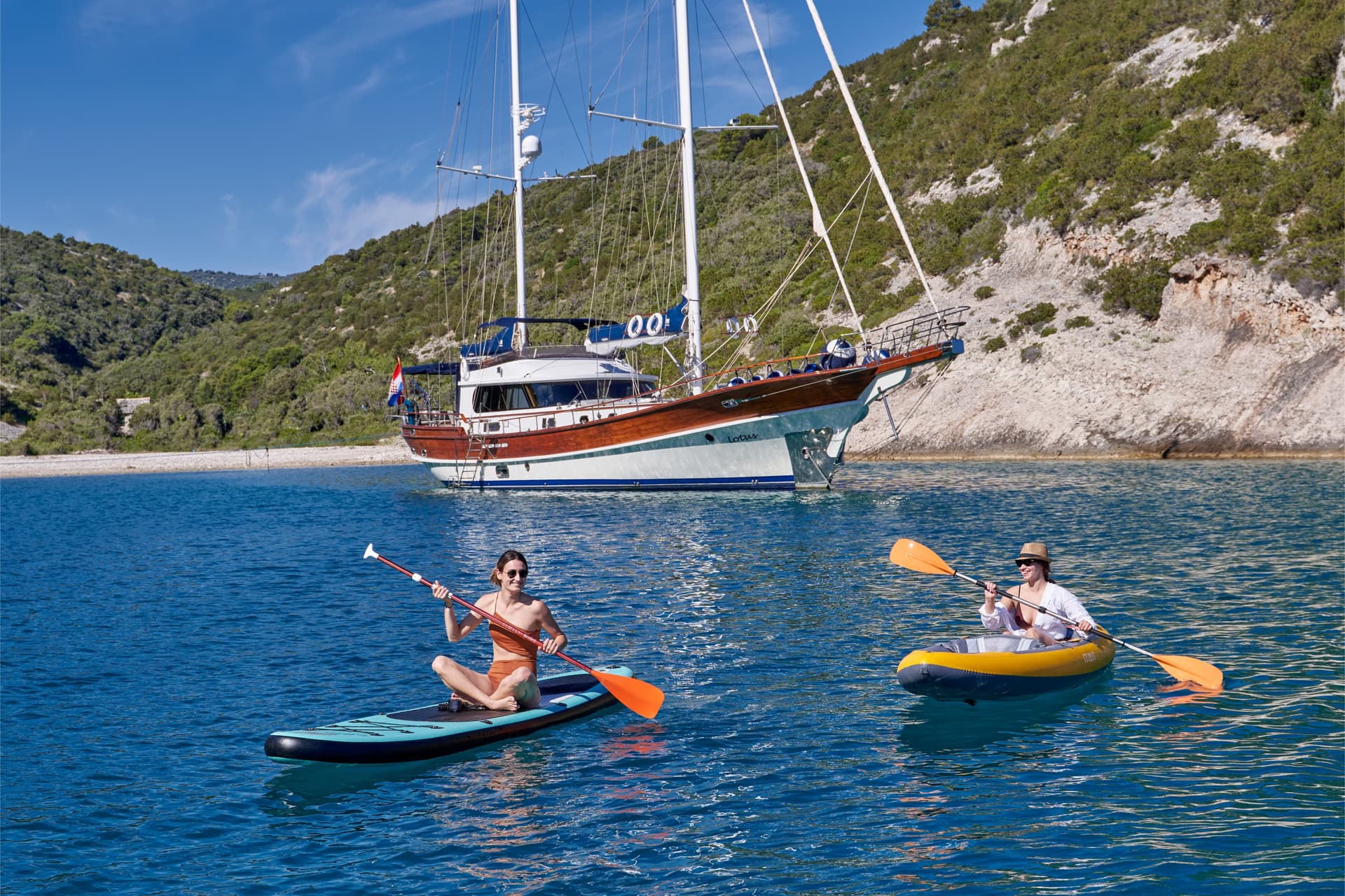 Paddleboarding and kayaking on blue water near a large sailboat and green coastline.