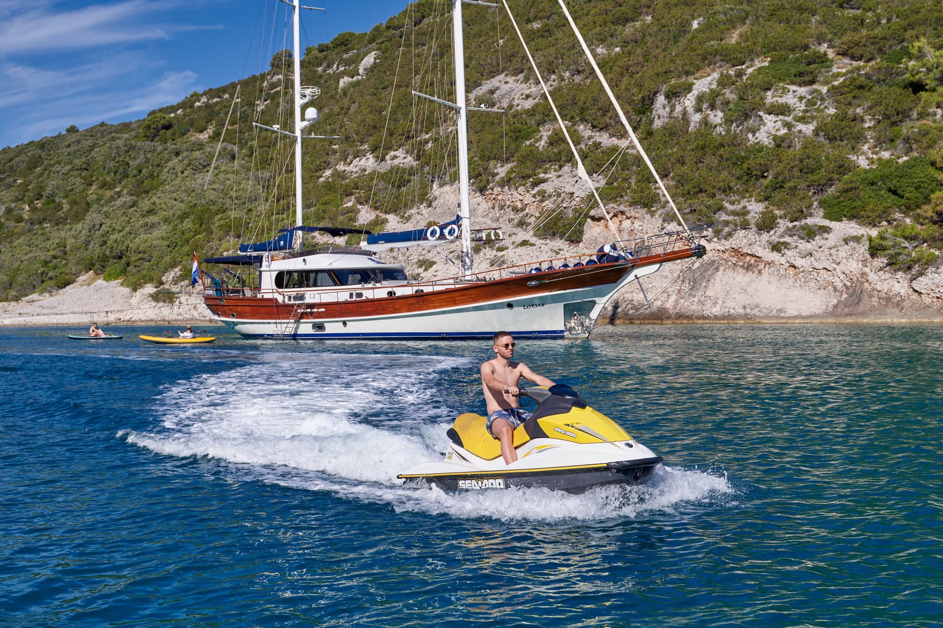 Man riding yellow Sea-Doo jet ski near moored wooden sailboat by rocky, green coastline.