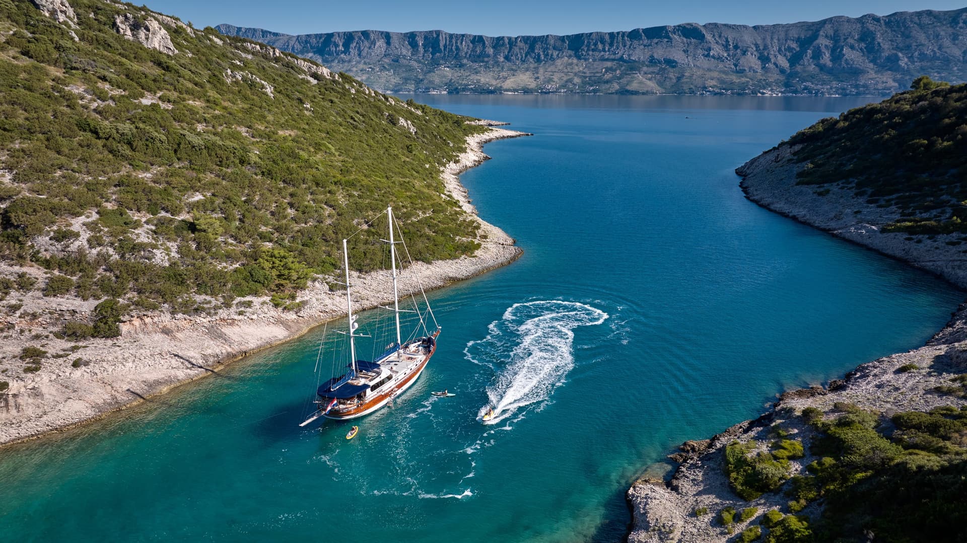 Large sailboat anchored in turquoise cove between steep, scrub-covered hills with distant mountains.