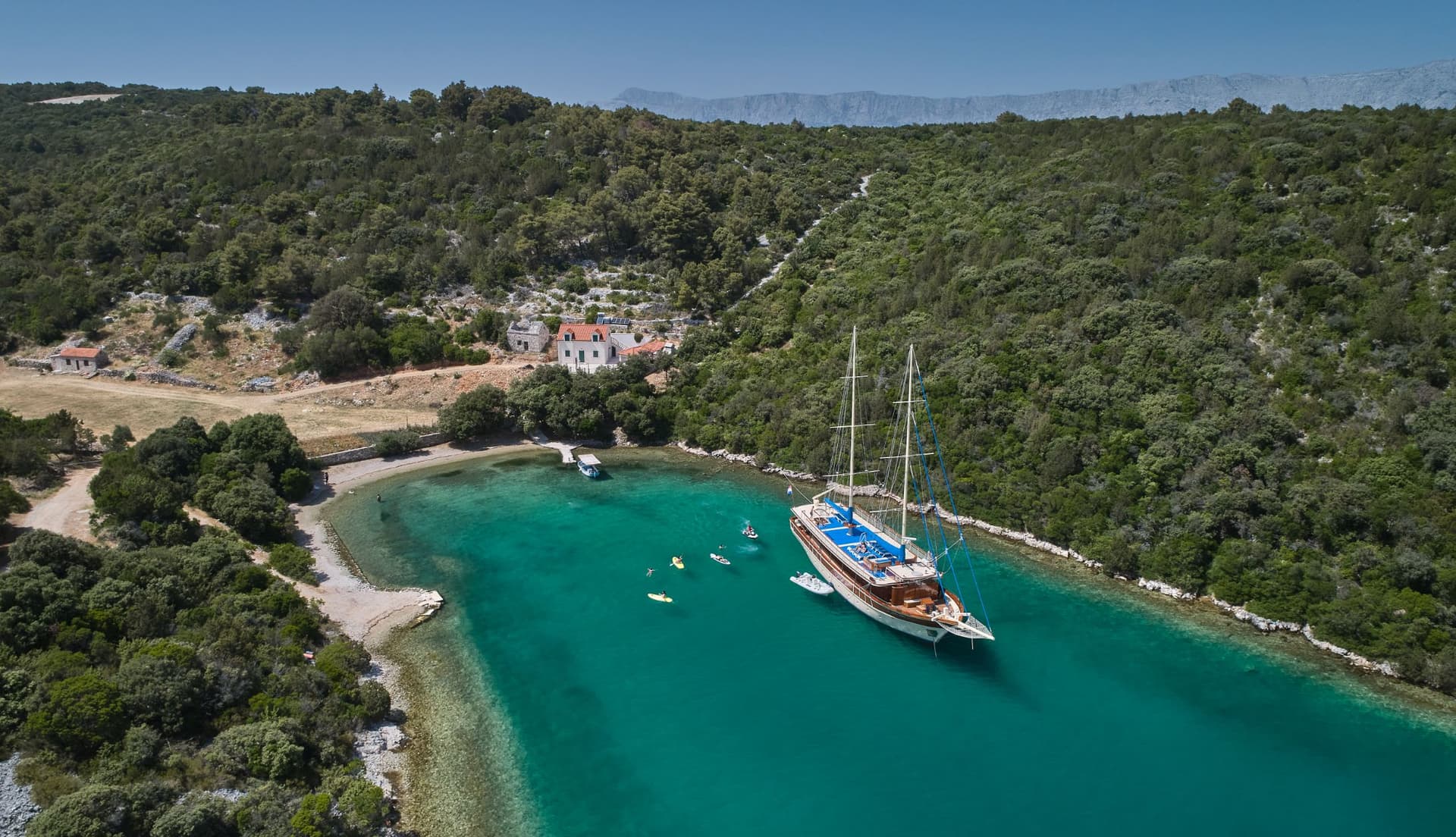 Large sailboat anchored in turquoise cove with paddleboarders near forested coastline.