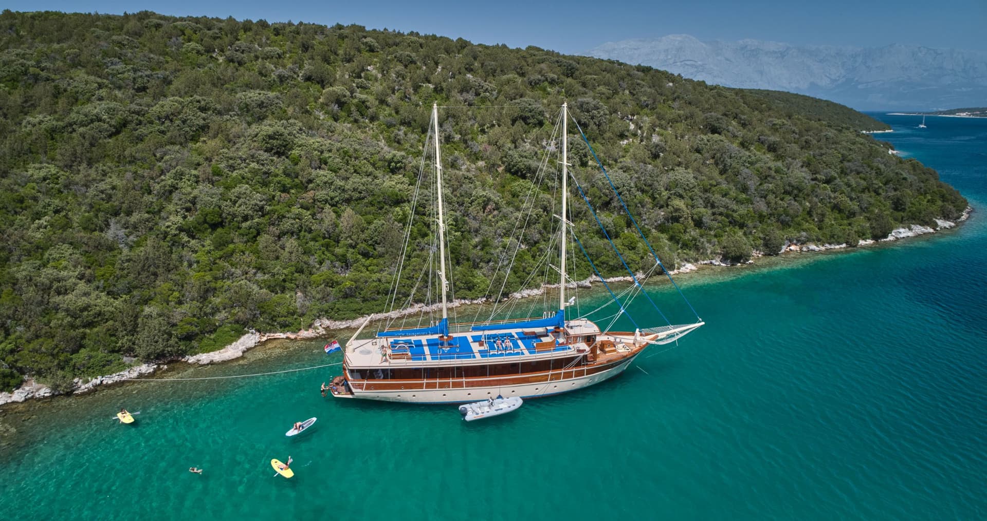 Large wooden sailboat anchored near forested Croatian coast with people paddleboarding in turquoise water.