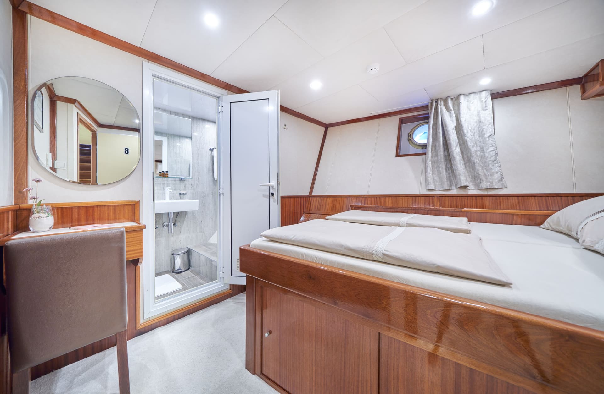 Cabin interior with double bed, ensuite shower, and porthole window on a boat.