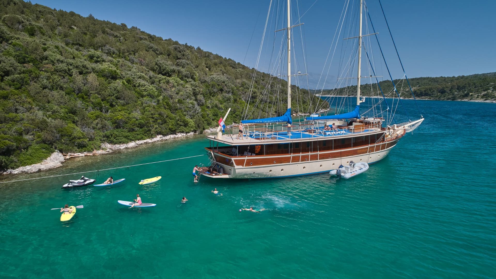 Large wooden sailboat anchored near a lush green coastline with people swimming and paddleboarding.