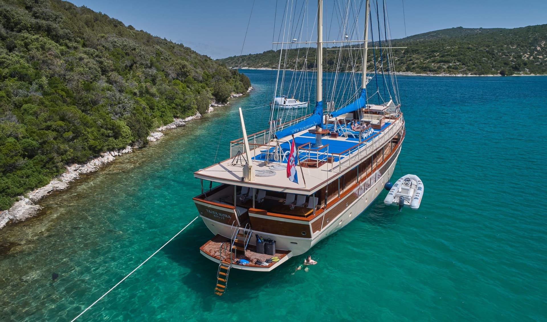 Large wooden sailing boat anchored in turquoise cove near forested coastline