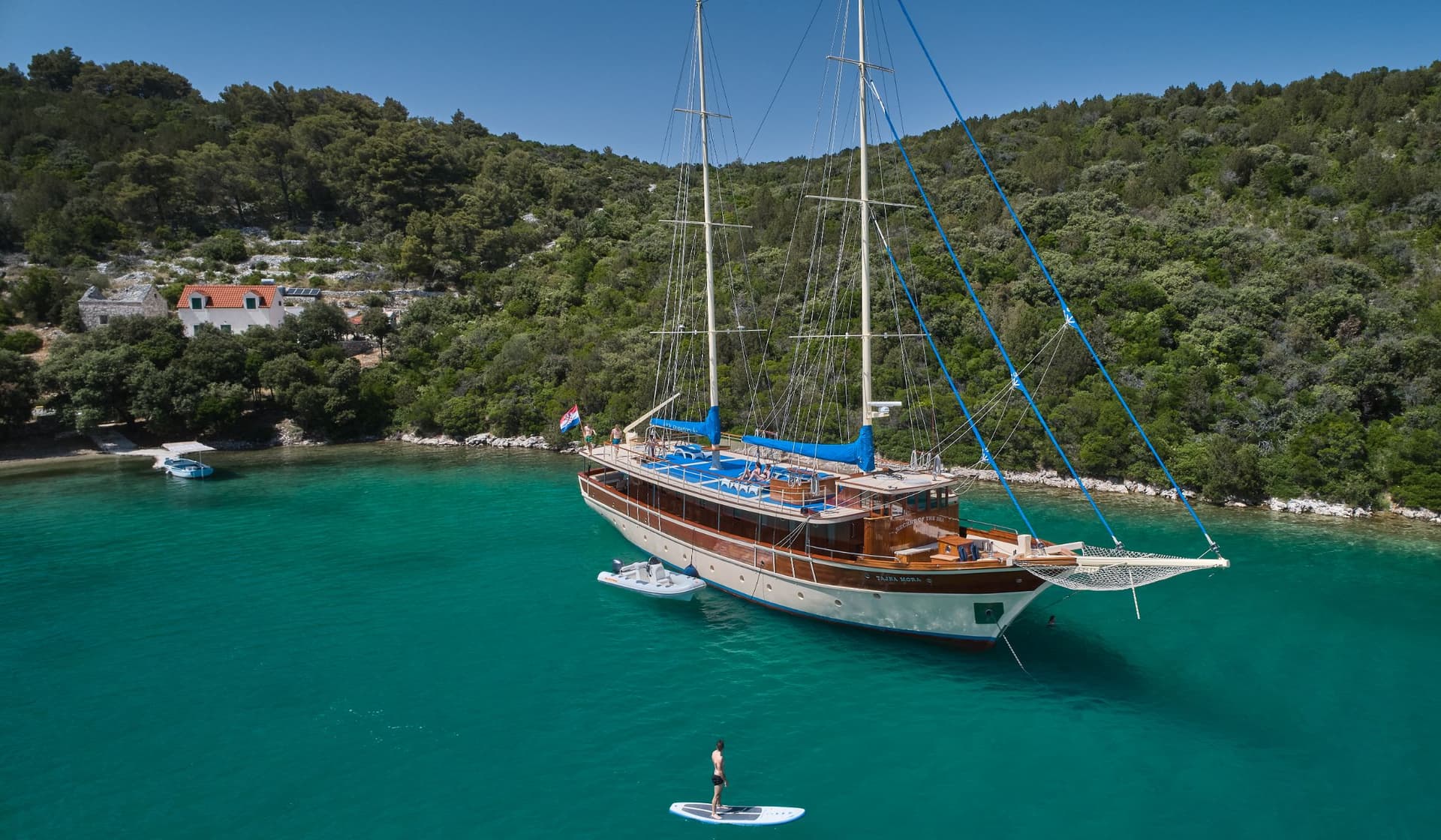 Large wooden sailboat anchored in turquoise cove near forested coast with person paddleboarding.