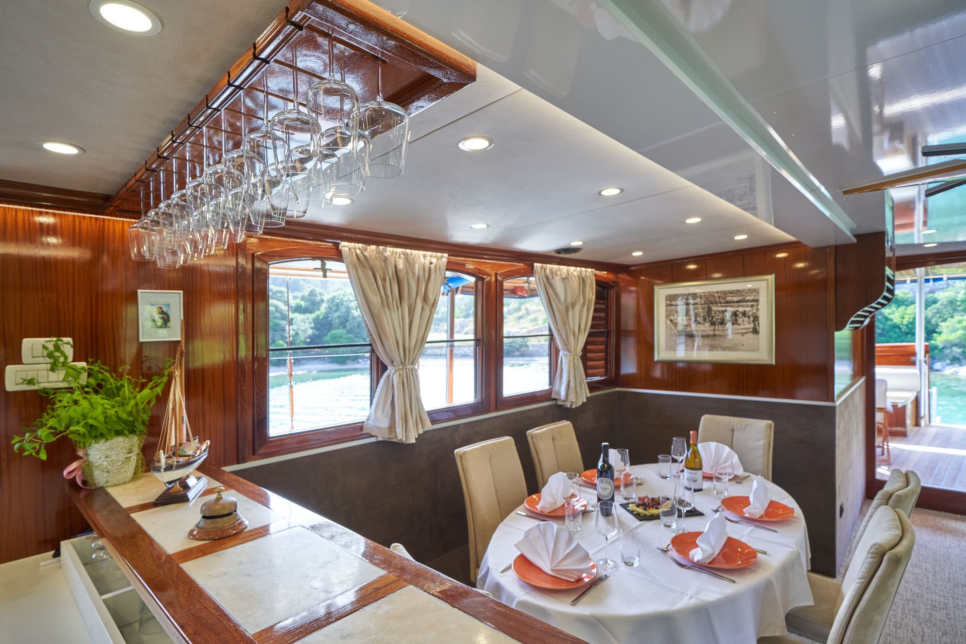 Indoor dining area on a boat with wood paneling and a set table overlooking green coastline.