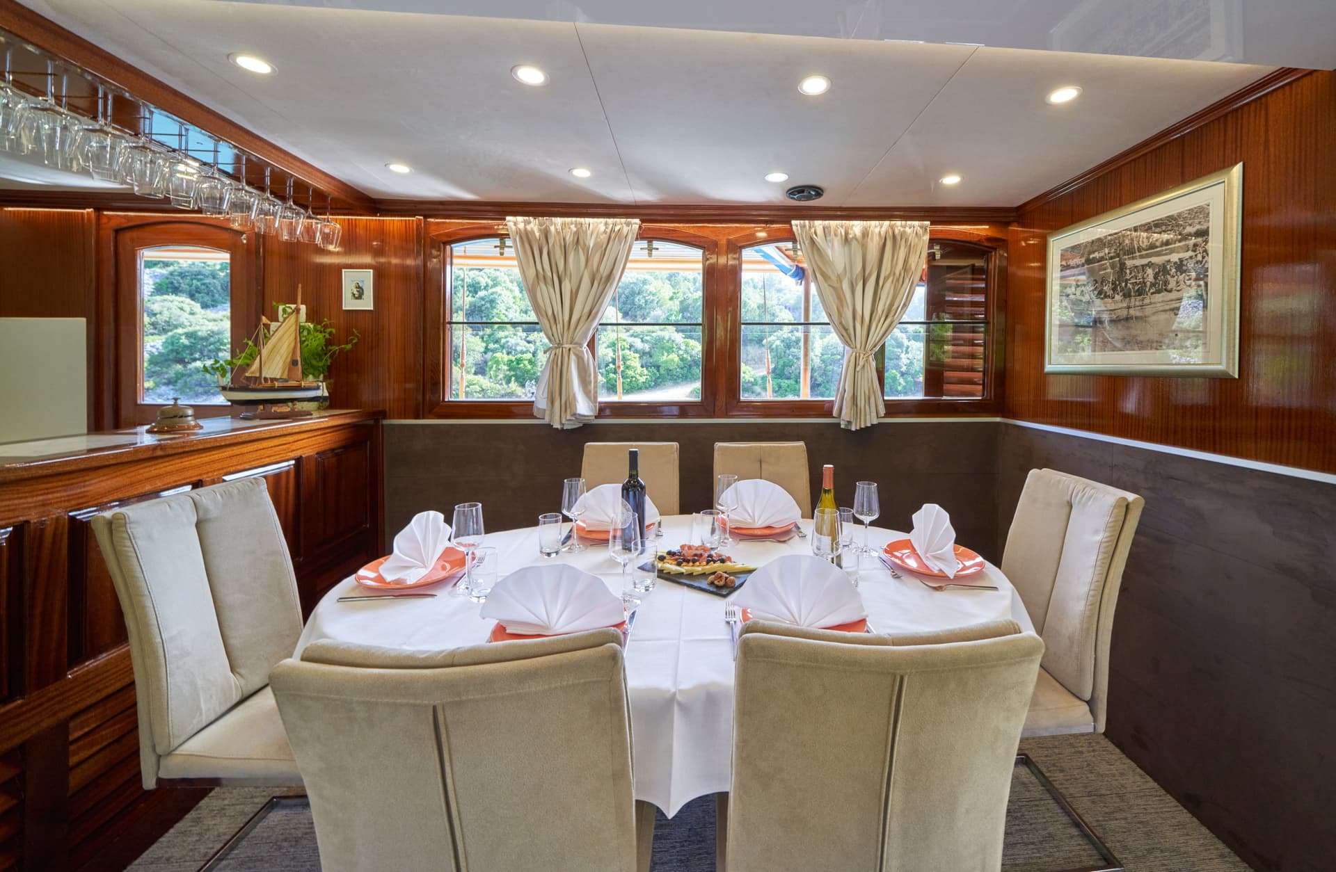 Dining table set for six inside a boat cabin with wood paneling and windows showing greenery.