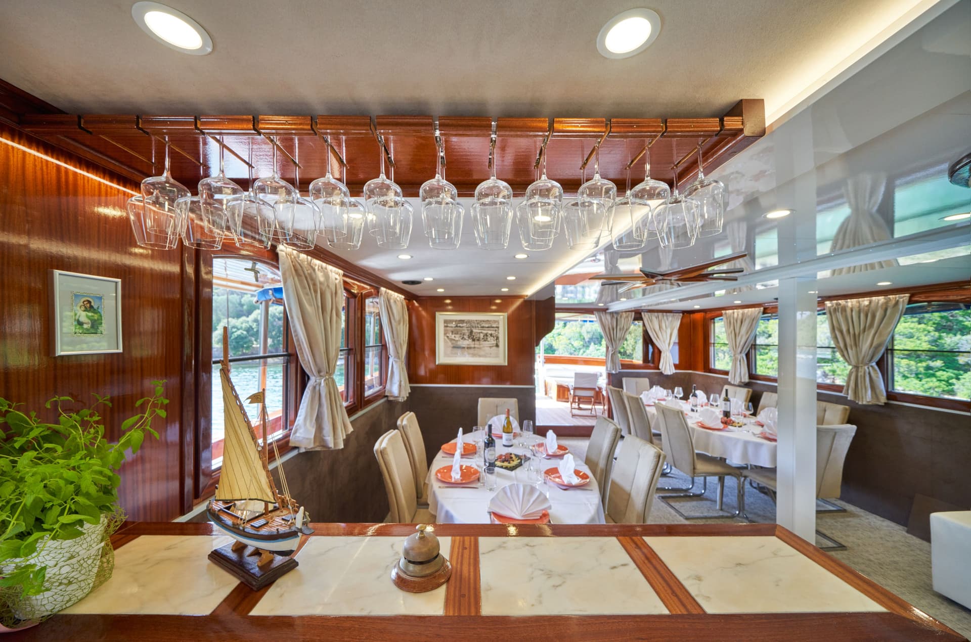 Dining room interior on a boat with wood paneling, set tables, and views of lush greenery outside.