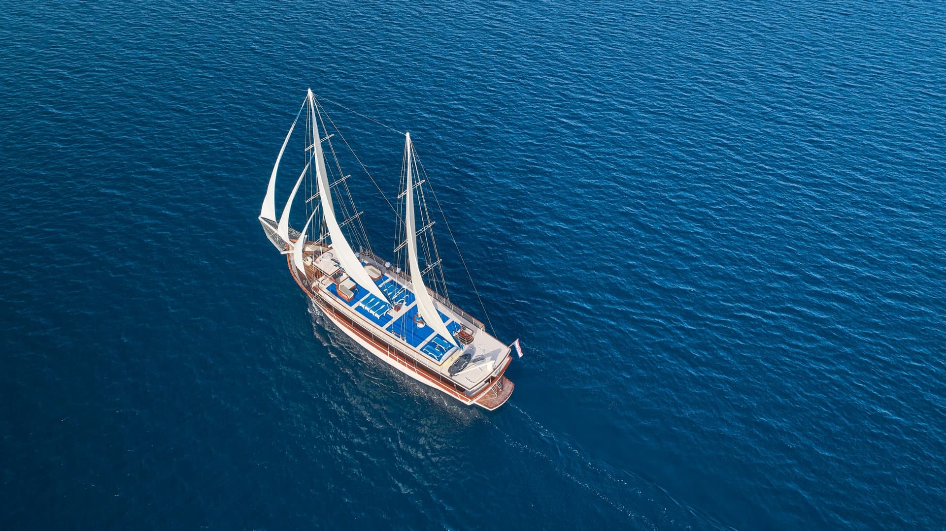 Two-masted sailboat with white sails cruising on deep blue, rippling sea water.