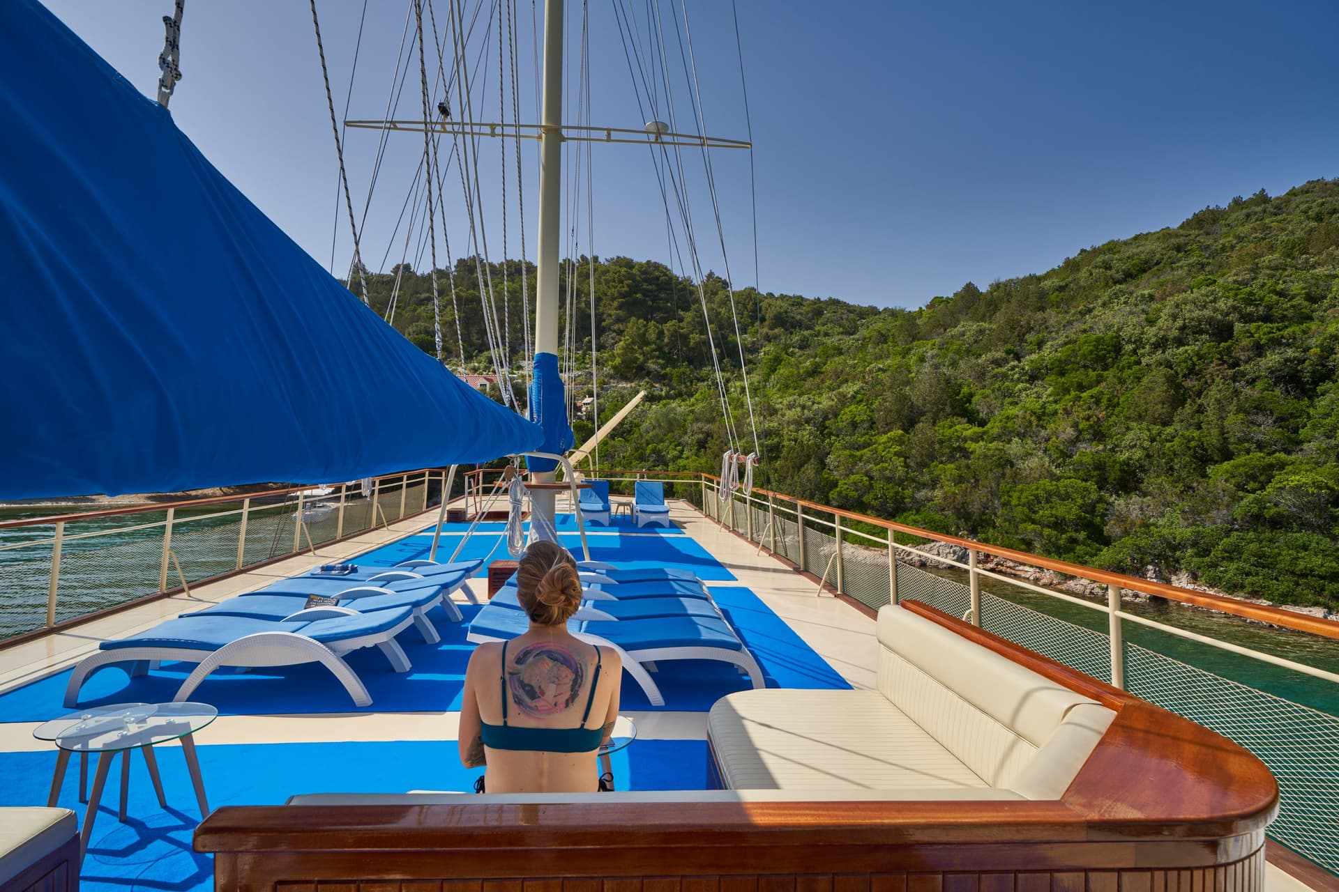 Woman relaxing on deck with blue sunshade near lush green coastline.