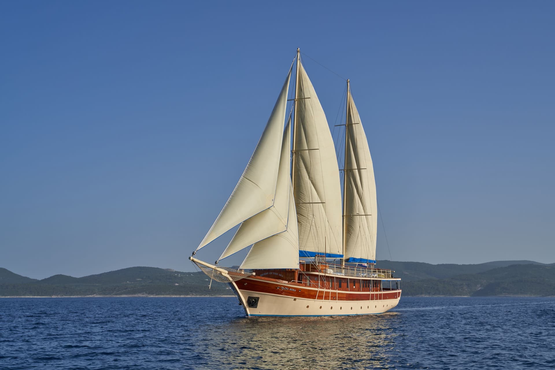 Wooden sailboat named Tajna Mora sailing on blue sea near forested coastline.