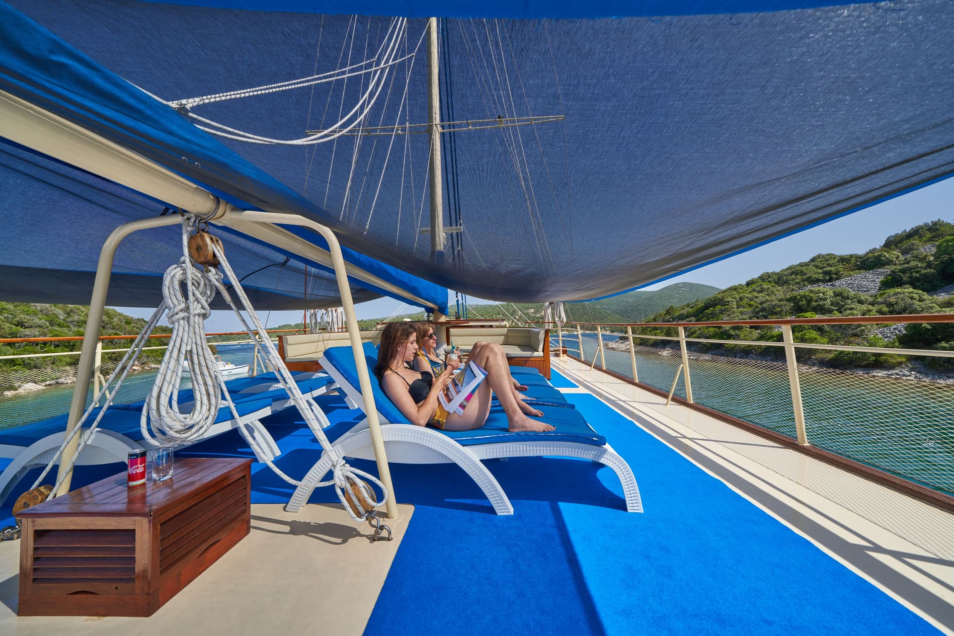 Women sunbathing on deck chairs under a blue awning on a boat near a green coastline.