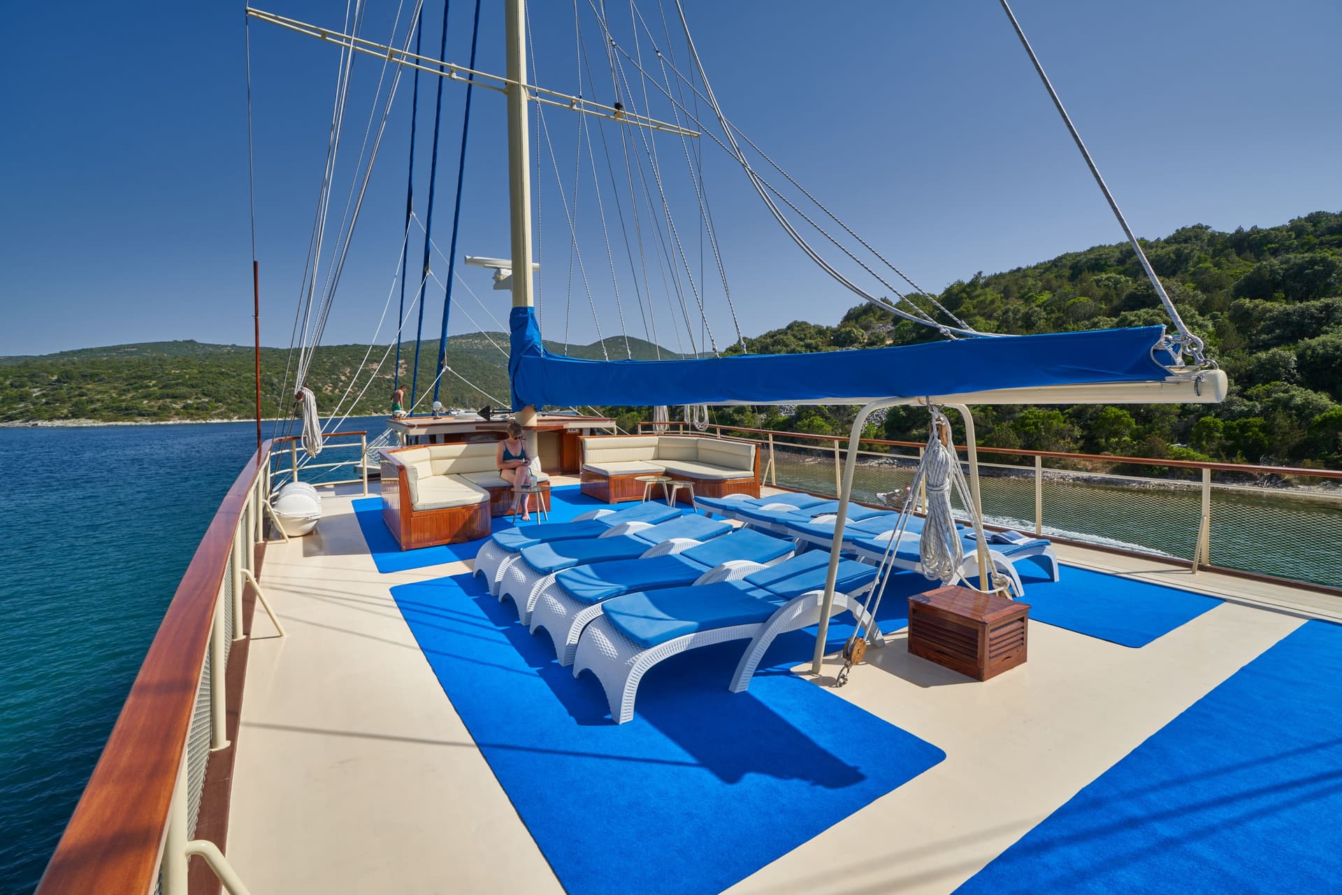 Sundeck of a boat with blue lounge chairs, seating, and mast rigging near a forested coastline.
