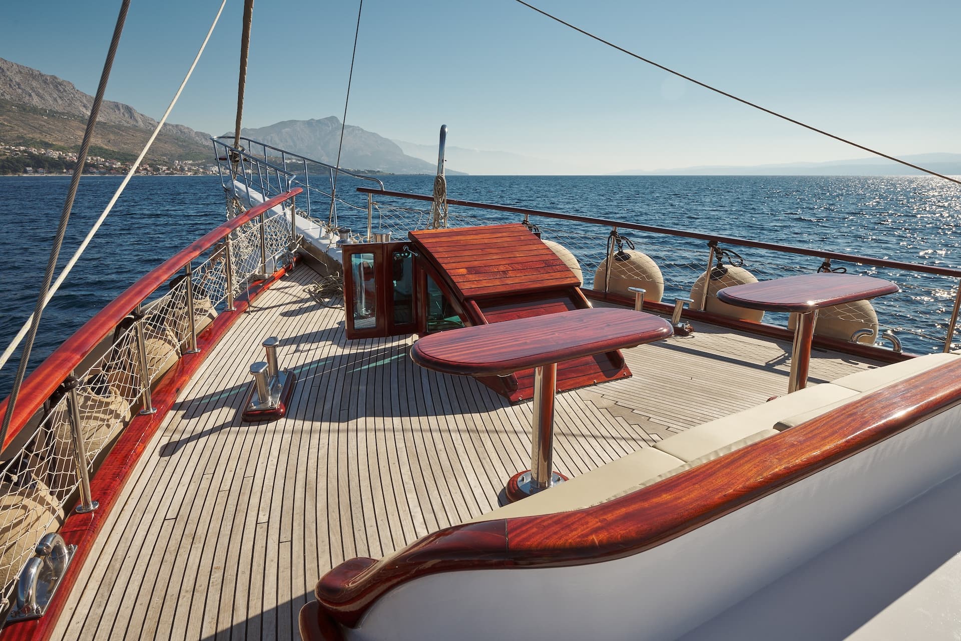 Wooden deck of a yacht with polished tables sailing near a mountainous coastline.