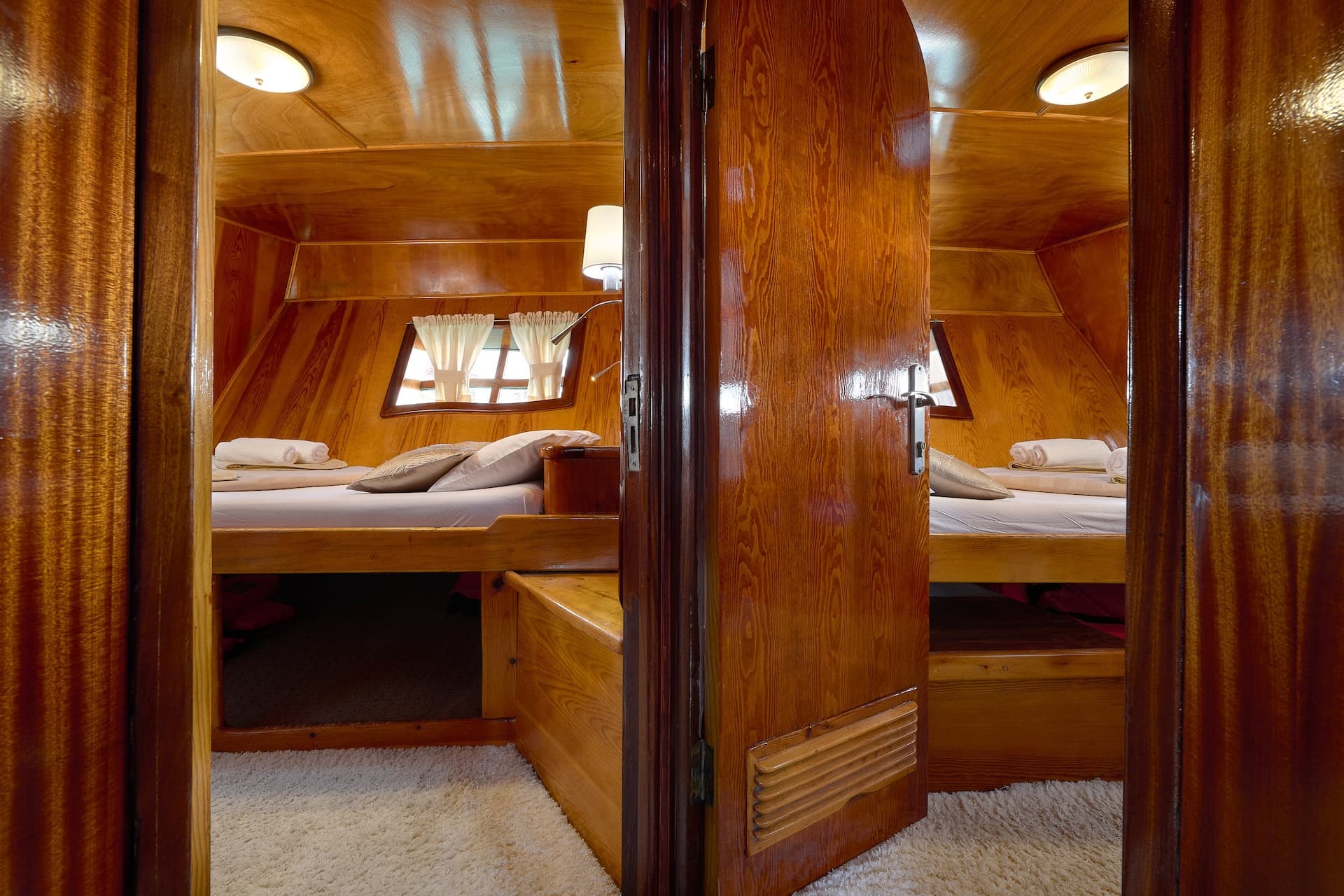View into a cabin with glossy wood paneling, twin beds, and white carpet.