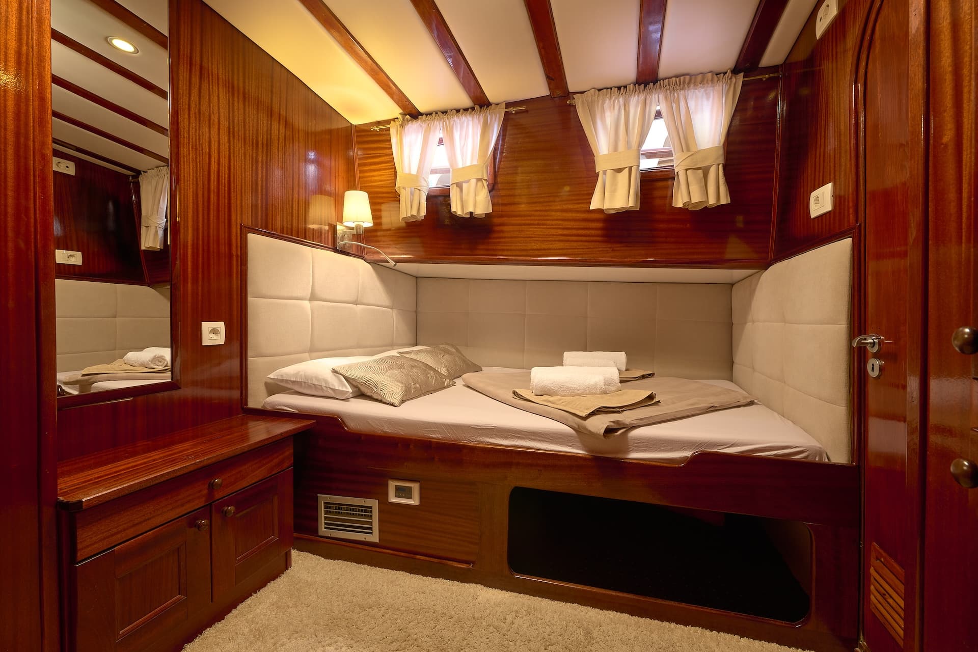 Cabin with double bed, wood paneling, and small windows with curtains, likely on a boat.
