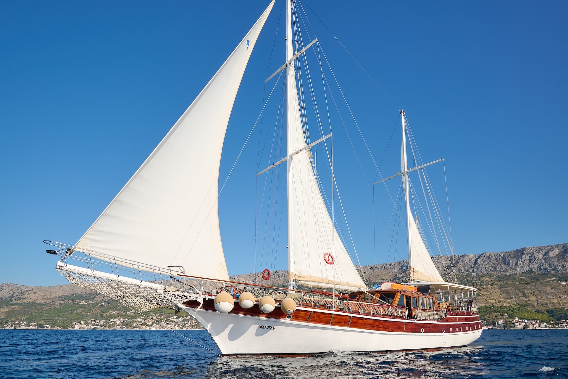 Wooden sailboat named Linda with full white sails on blue sea near coastal town and mountains.