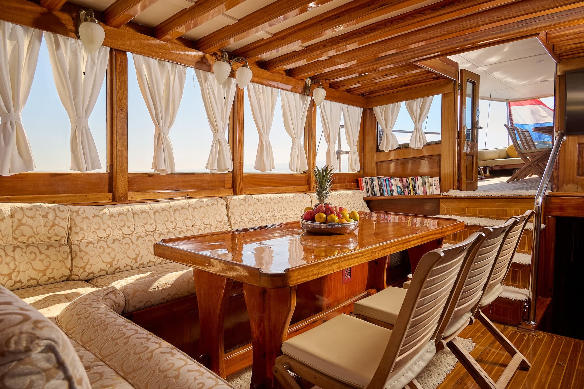 Interior of wooden yacht salon with dining table, fruit bowl, and view of deck.