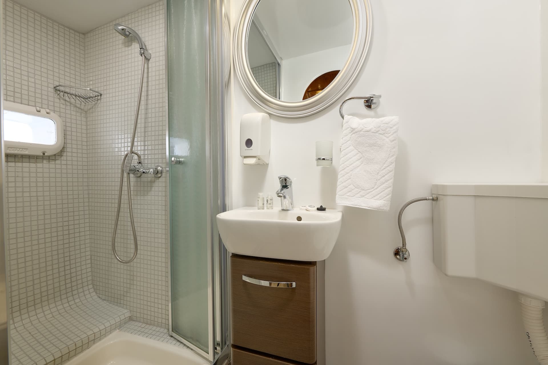 Compact bathroom with white tiled shower, small sink vanity, and round mirror. Porthole window visible.