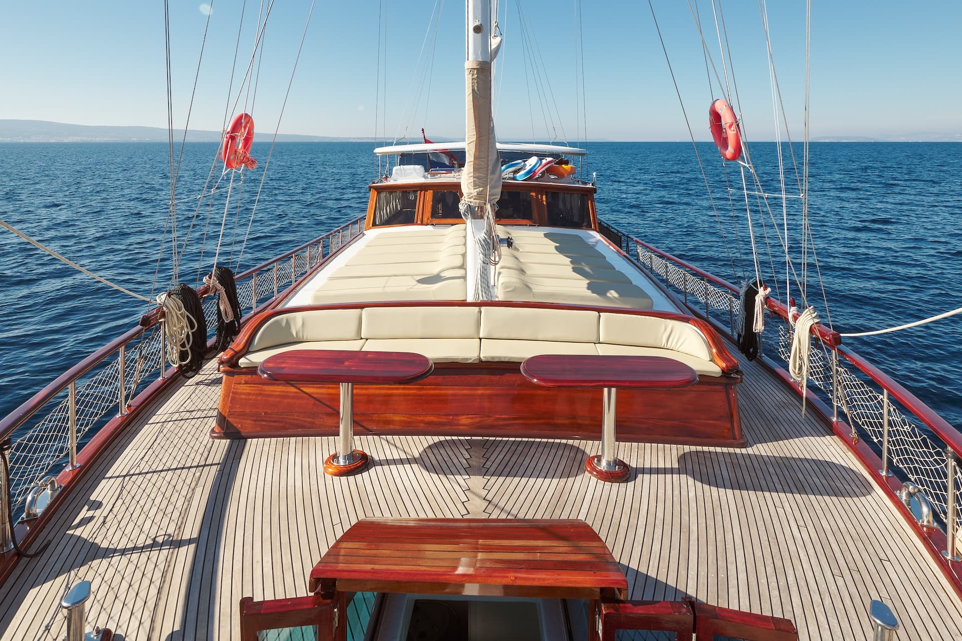 Wooden deck seating area on a boat sailing on deep blue water with distant coastline.