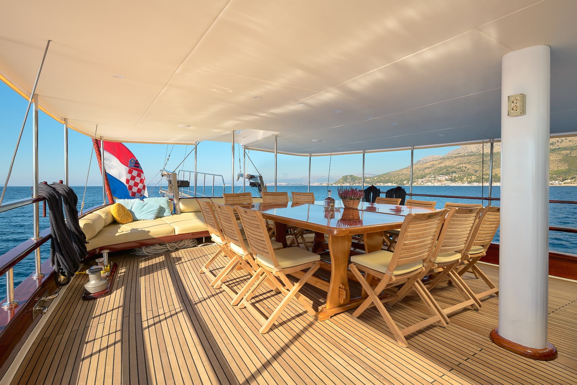 Outdoor dining area on a yacht with teak deck, Croatian flag, and coastal mountains.