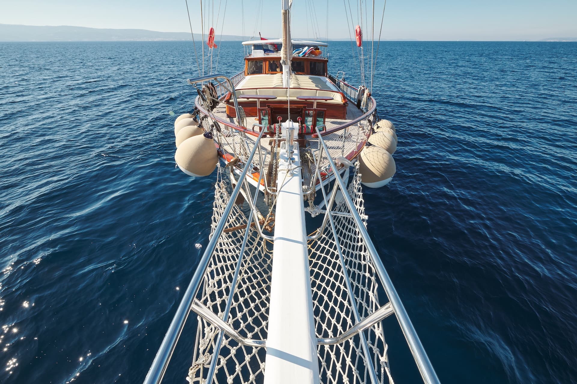 View from bowsprit netting onto wooden boat deck sailing on deep blue sea toward distant coast.