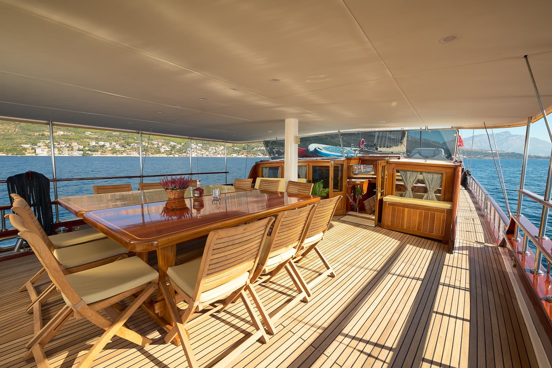 Outdoor dining area on a yacht deck with wooden table, chairs, and coastal view.