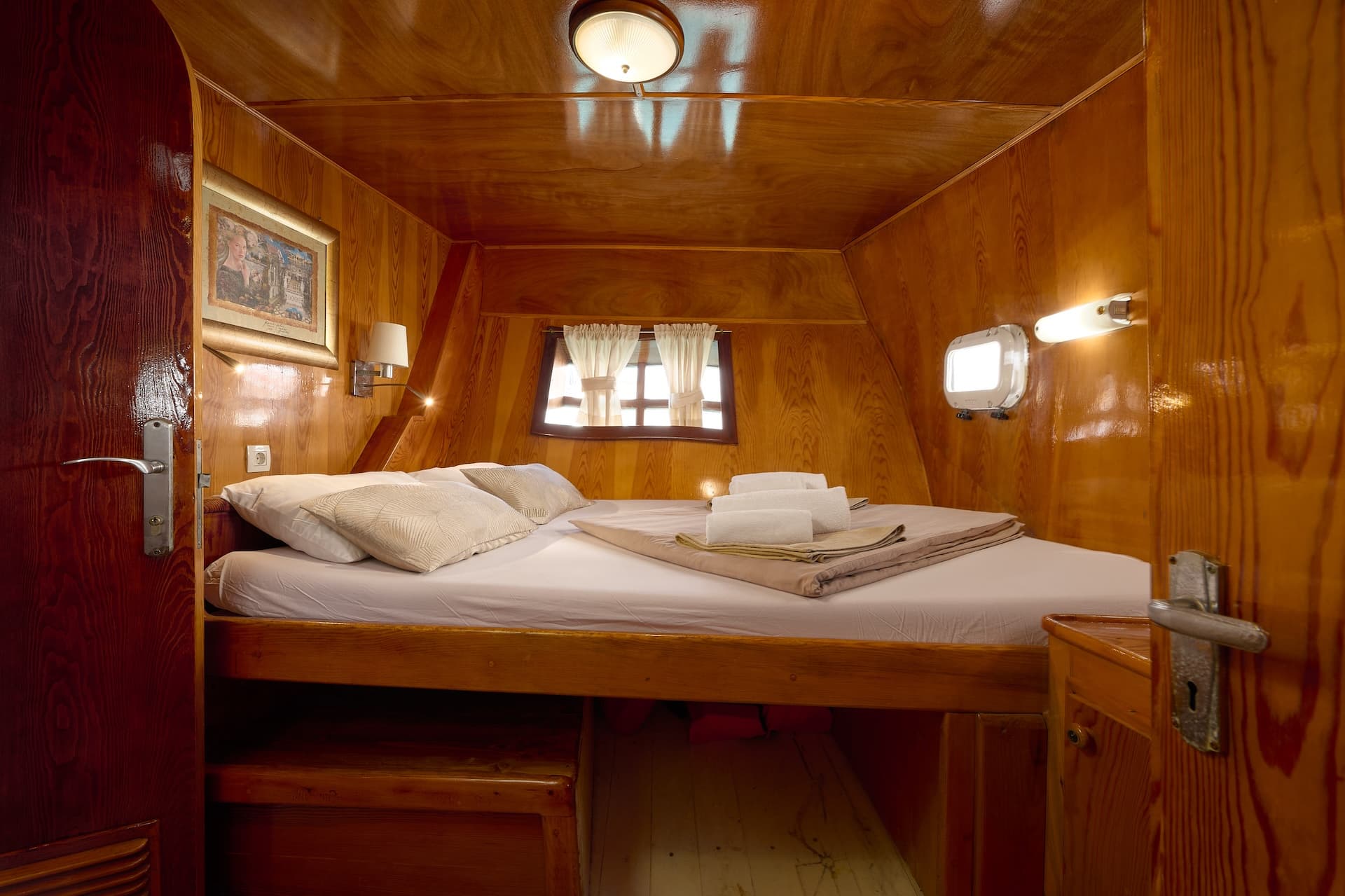 Cabin bedroom with wood paneling, double bed, and porthole window, likely on a boat.