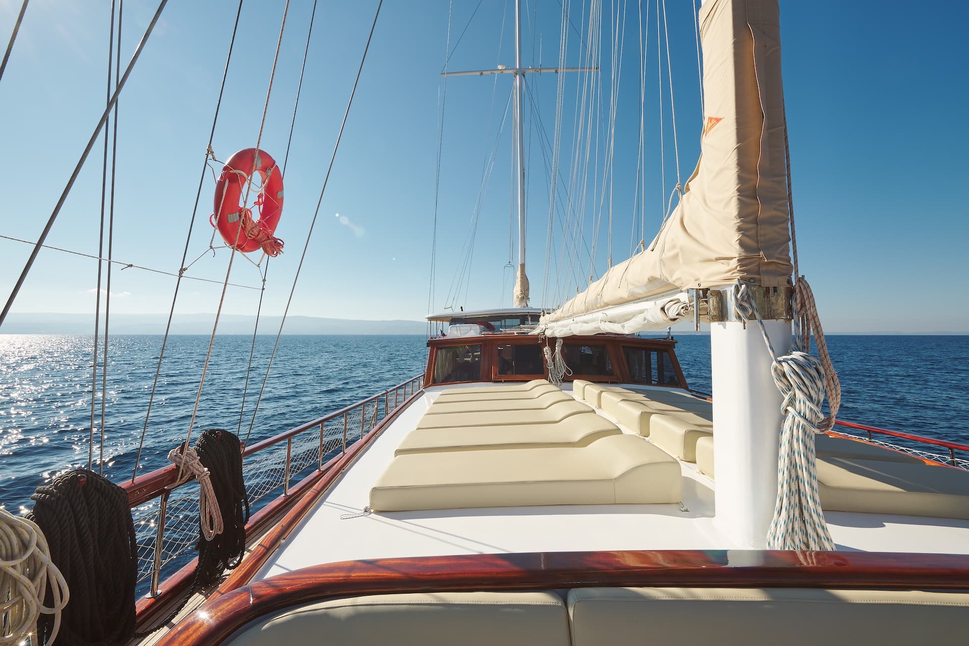 Deck of a sailboat with sunbeds, ropes, and a life preserver on the water.