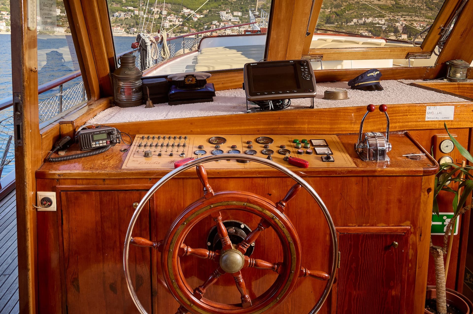 Boat helm with wooden wheel, gauges, and captain's hat overlooking a coastal town.