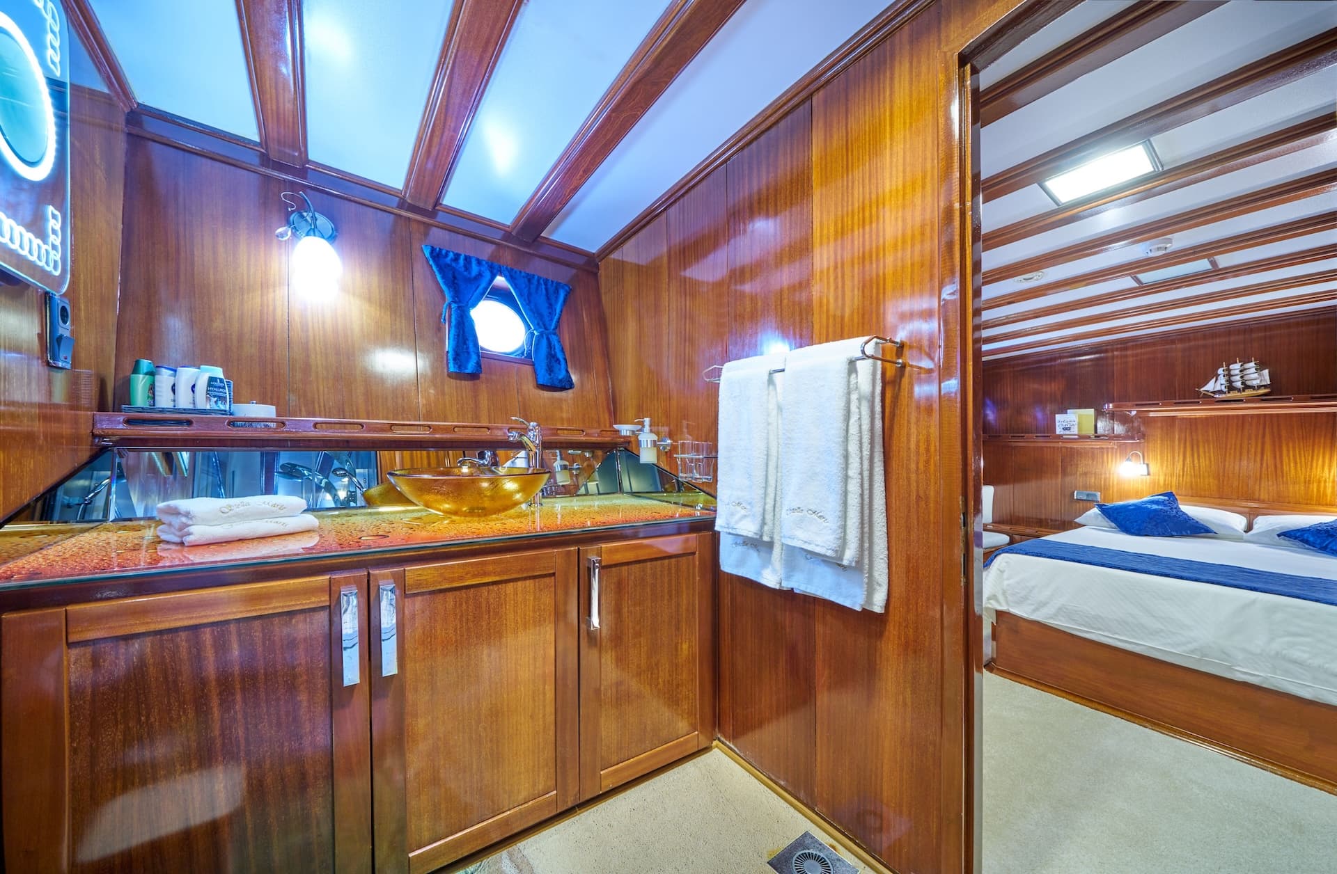 Yacht bathroom with wood paneling, glass sink, and view into a cabin with a bed.