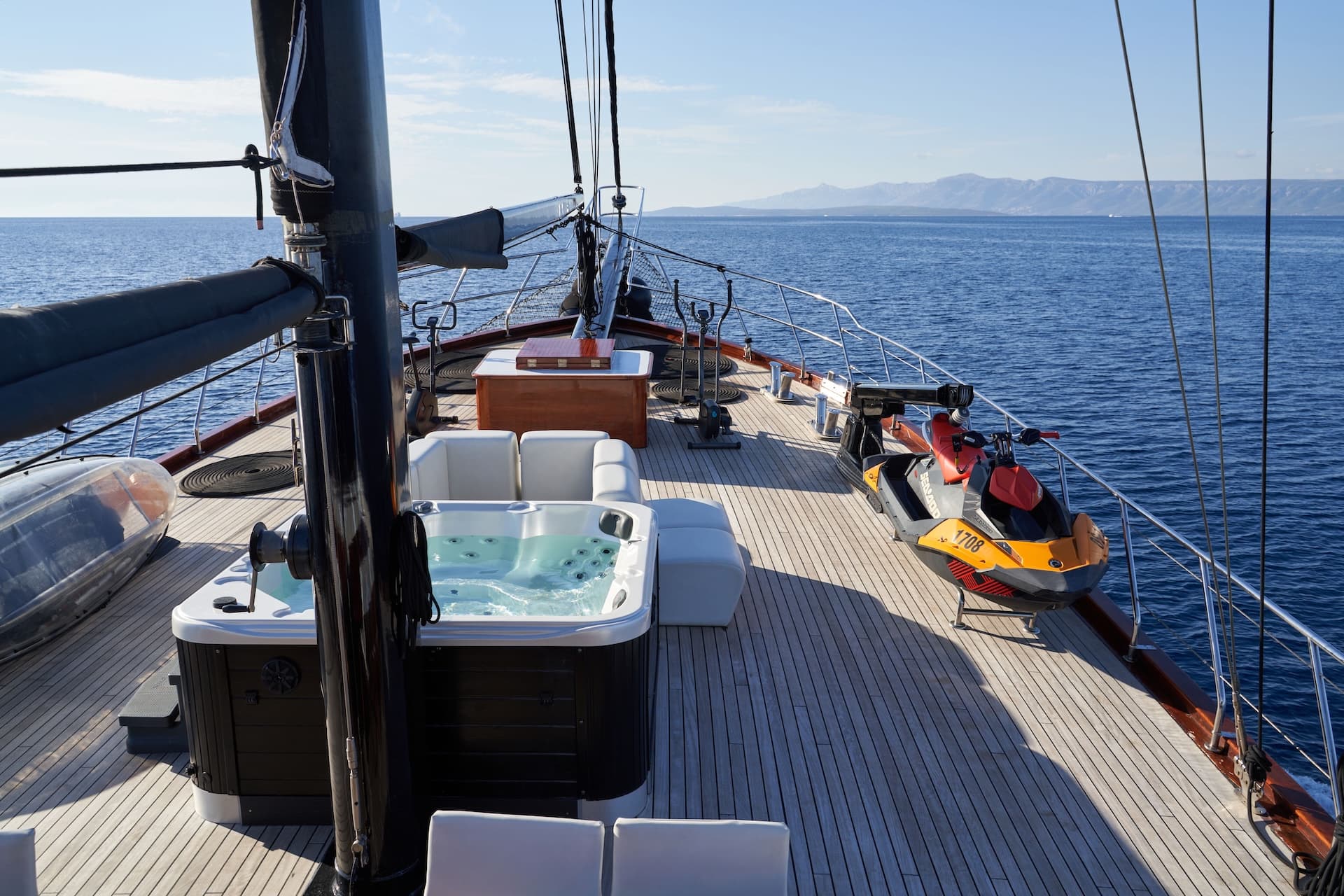 Deck of sailboat with jacuzzi, jet ski, and mountains visible across the blue sea.