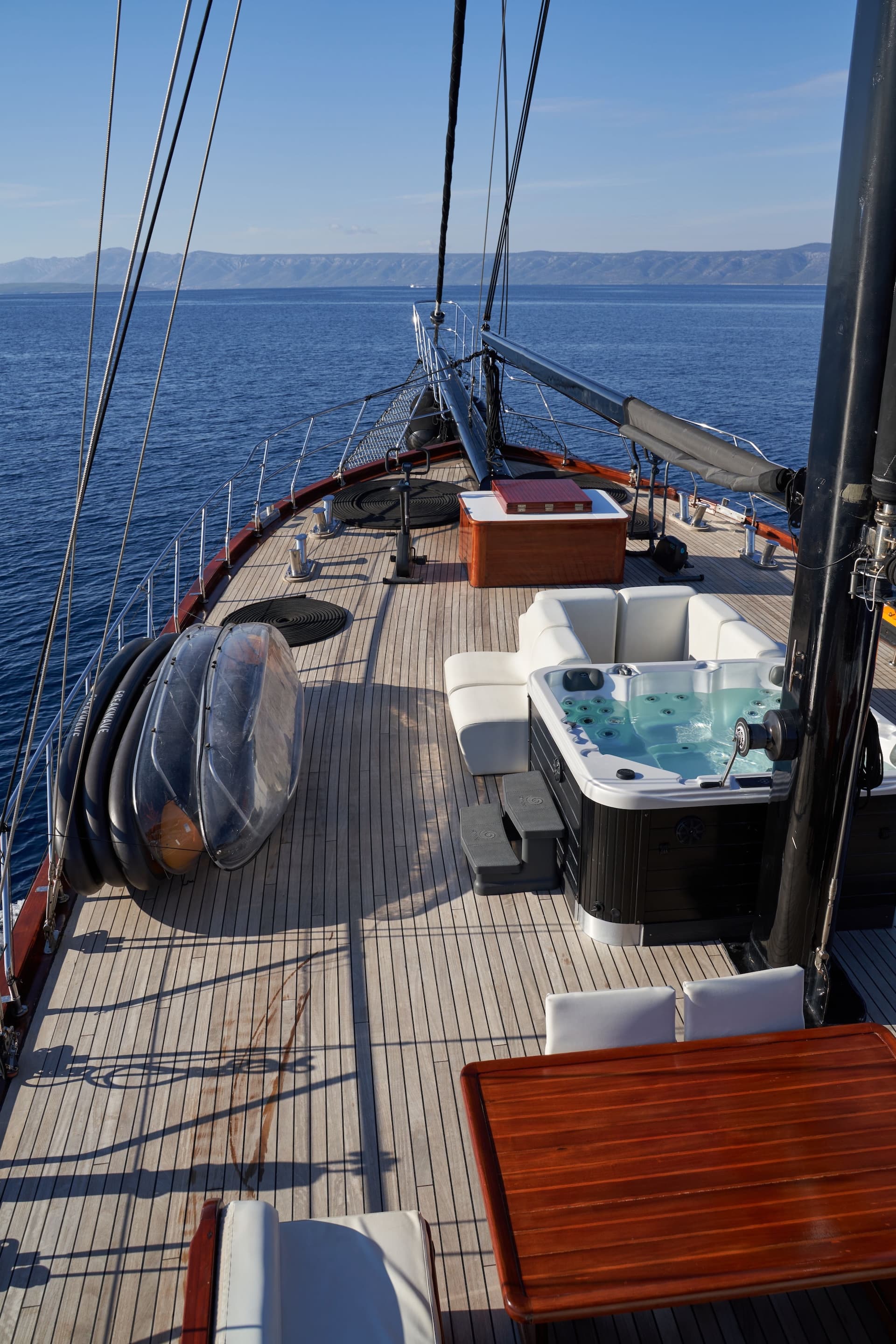 Bow deck of a yacht with a hot tub, exercise bike, and wooden table, facing mountains across the sea.